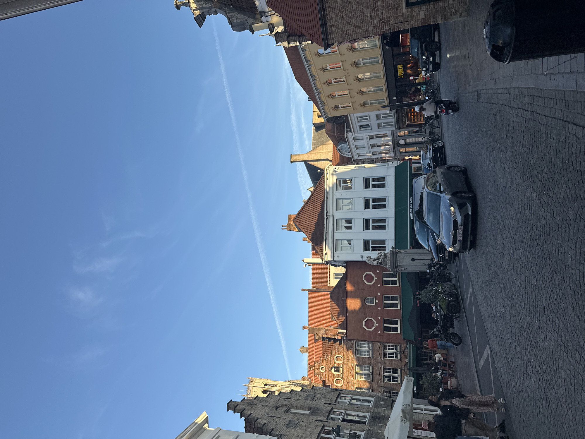 The image depicts a street scene in Bruges, Belgium, likely Market Square or a similar central area, given the characteristic architecture. The sky is a clear blue with faint white contrails from an aircraft. The buildings are a mix of historical European styles, with red-tiled roofs and varied facades. On the left, a tall bell tower, possibly part of the Belfry of Bruges, is visible. Several cars, including a silver hatchback and a black Jeep, are on the cobblestone street. A person is riding a scooter in the middle of the road. In the foreground, two people stand on the left, and a black trash bin is on the right. Outdoor seating for a cafe or restaurant is visible on the left. The overall impression is a bustling urban environment under a bright sky.