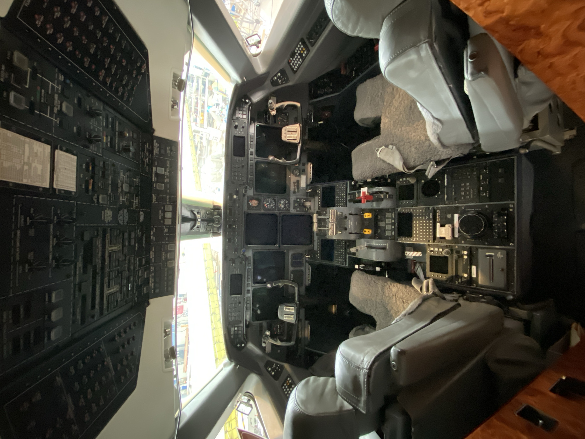 The image depicts the interior of an aircraft cockpit, specifically the area containing the instrument panels, seats, and forward visibility. The left-hand panel is densely populated with various switches, buttons, and instruments, while the center panel features multiple display screens and control interfaces. Two pilot seats are visible, upholstered in light gray leather and positioned in front of the instrument panels. The cockpit's windows offer a view of the exterior environment, where a structure with yellow elements is visible. The image was likely taken indoors, possibly within an aircraft hangar or maintenance facility. Given the prompt, it is implied that this location is in Skulte, Latvia.