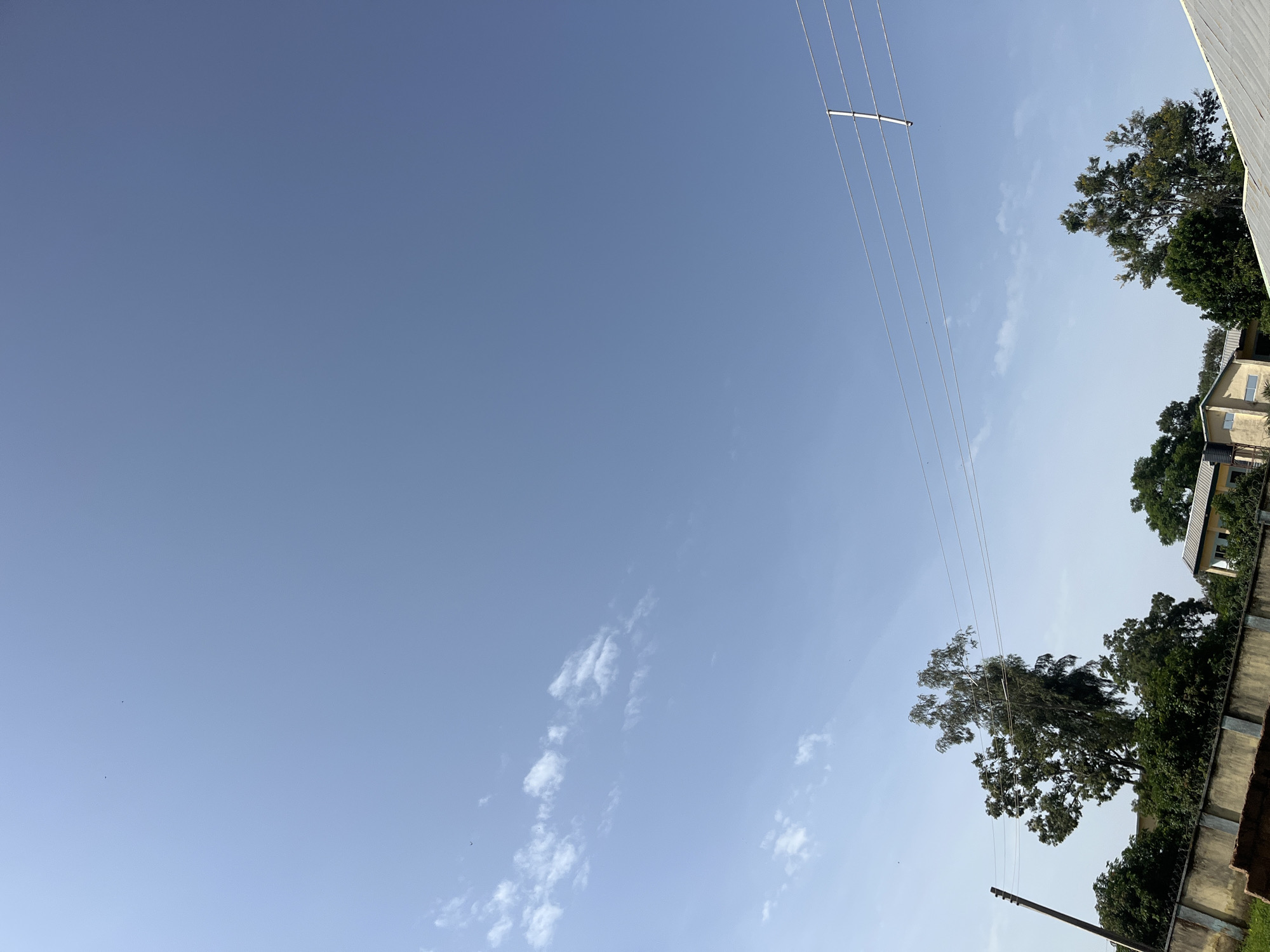 The image depicts a clear blue sky with scattered white clouds, indicating fair weather. A power line runs diagonally across the frame from the upper right to the lower left, supported by a dark pole visible in the lower center. To the right, several trees with lush green foliage are present, along with the corner of a building with light-colored walls and a beige-colored roof. A concrete wall with barbed wire runs along the bottom edge of the image, partially obscuring the view of the building and trees. The scene appears to be set outdoors, likely in a rural or suburban area, given the presence of trees, a building, and power lines. The lighting suggests it is daytime, possibly morning or afternoon. No people are visible in the image. There is no visible text. The overall impression is one of a serene, everyday scene in Bukuru, Nigeria.
