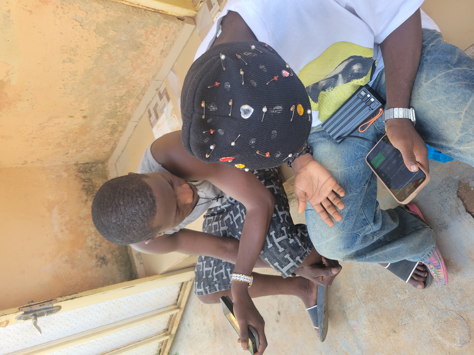 Two individuals are seated outdoors on a concrete step in front of a textured, weathered wall. The person on the left, wearing a grey tank top, patterned shorts, and dark sandals, holds a mobile phone in their left hand and wears a white and gold bracelet on their right wrist. They are looking down at the phone. The person on the right wears a black beanie adorned with various pins, a white t-shirt with a graphic print, and blue jeans. They are holding a mobile phone in their right hand and a black portable charger is visible in their lap. Their left hand is open, palm up. The environment suggests an urban or semi-urban setting with a metal gate visible to the left.