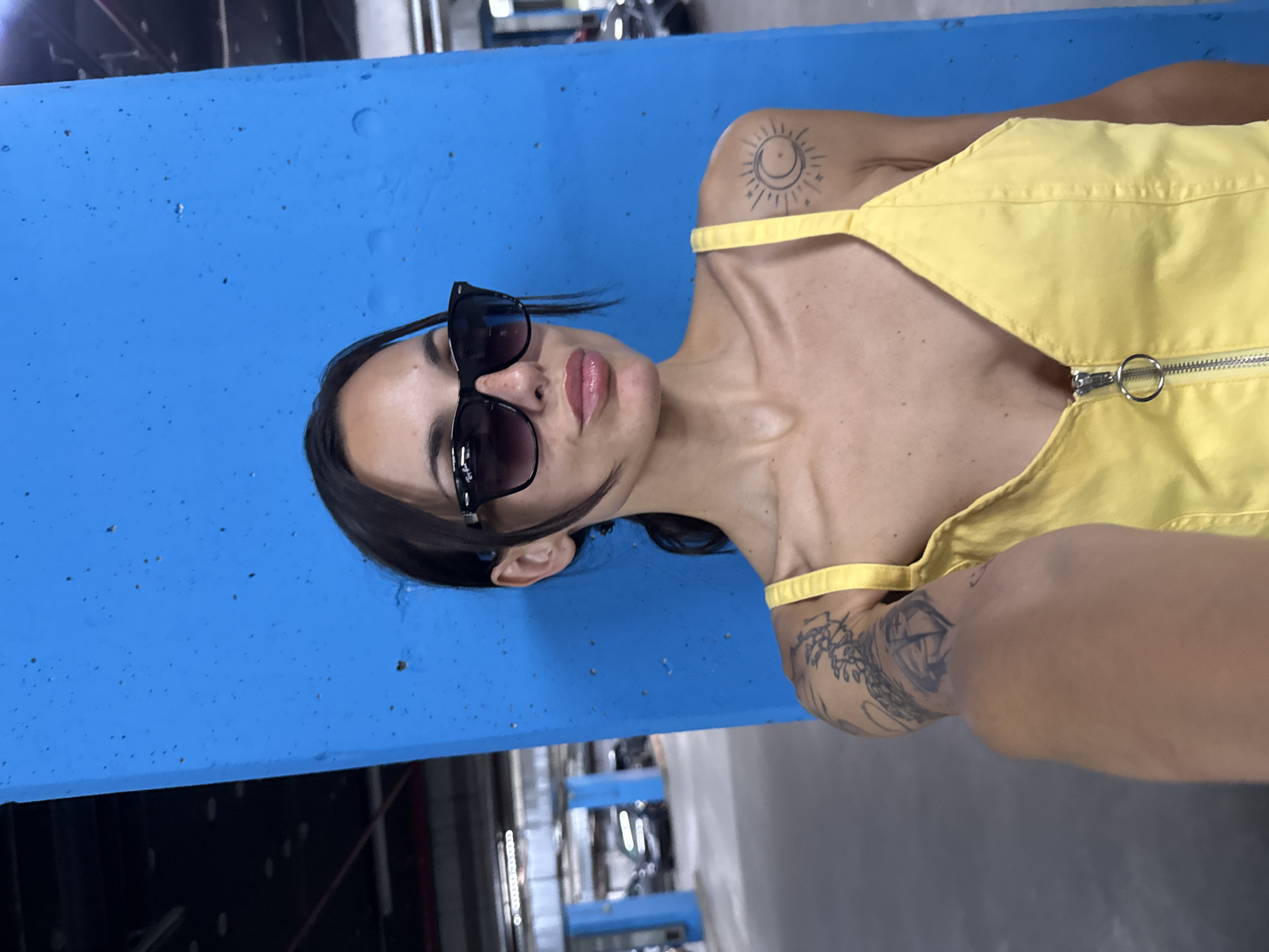 The rotated image captures a close-up selfie of a person, likely female, in Pleven, Bulgaria. The subject looks directly at the camera with a neutral expression, her lips slightly parted. She has dark hair pulled back and wears black sunglasses with "Ray-Ban" text visible on the arm.
She is dressed in a bright yellow garment, possibly an overall or dress, featuring a silver zipper with a circular ring pull on the front. Notable body art includes a stylized black ink tattoo of a sun and crescent moon on her right shoulder, and a larger, intricate black tattoo covering a portion of her left arm.
The immediate background on the left side of the frame is a vibrant, textured blue surface, resembling a concrete wall or beam, adorned with small dark specks. In the lower background, an indistinct, darker area suggests an indoor industrial or garage-like setting with glimpses of metallic structures. The lighting appears artificial, complementing the indoor environment.