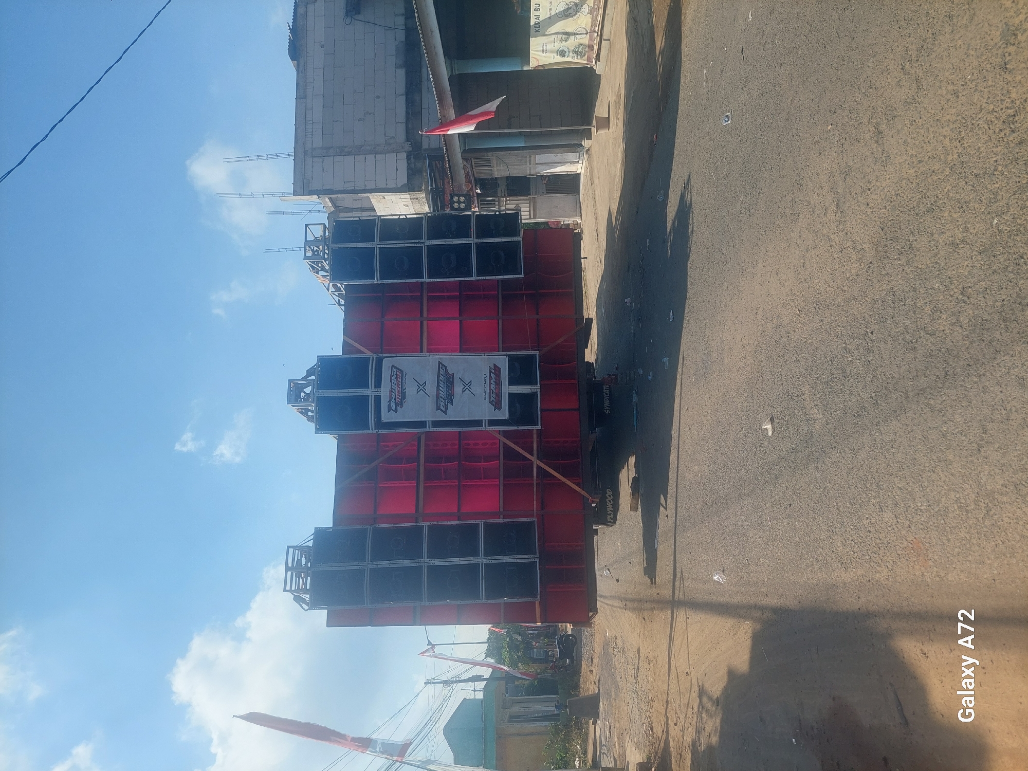 A large sound system is mounted on the back of a truck. The sound system is comprised of numerous speakers arranged in a grid pattern, with red panels between them. The truck is on a paved road in a somewhat rural or suburban area. The sky is blue with some clouds. Buildings and trees are visible in the background. A red and white flag is flying from a pole to the left of the truck. The scene appears to be in Sengguruh, Indonesia, as indicated by the presence of Indonesian flags and architecture. The overall impression is one of a mobile entertainment setup, possibly for an event or festival.