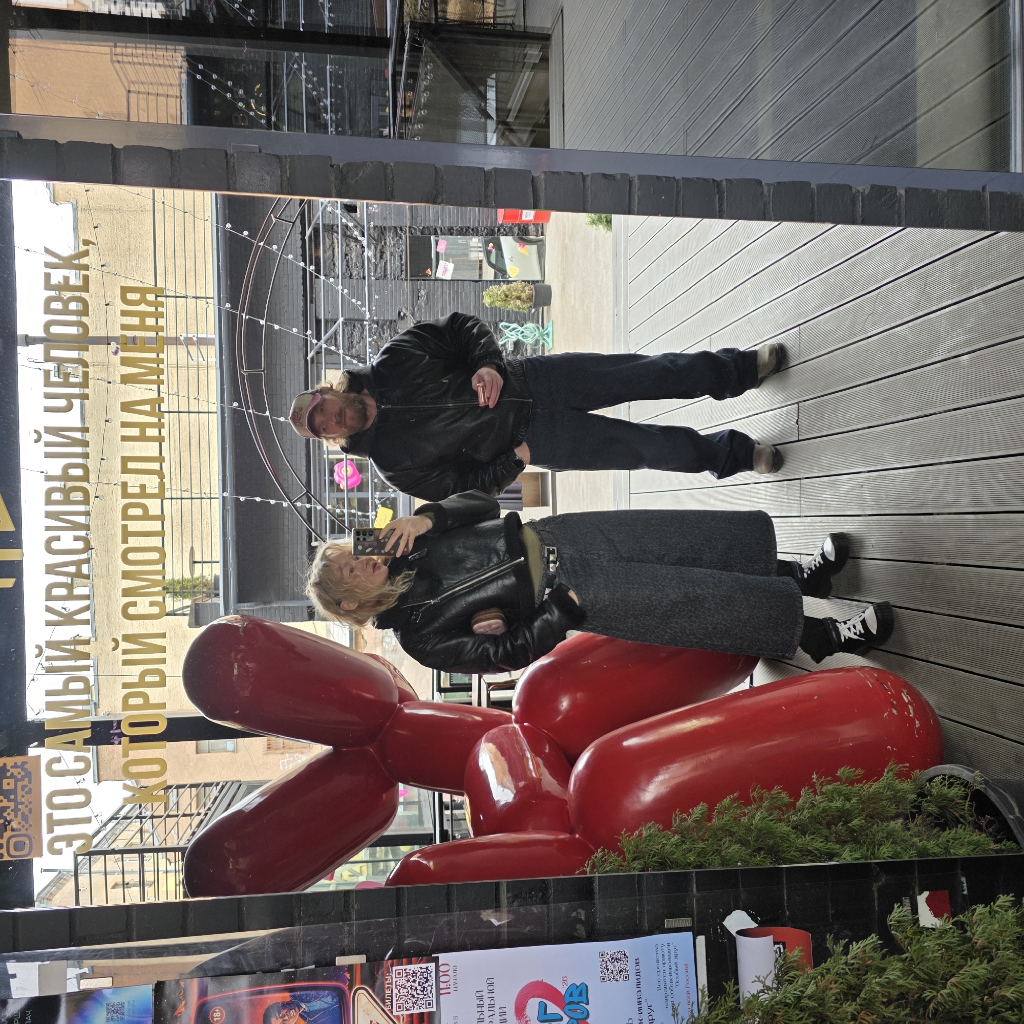 A portrait-oriented image, rotated 90 degrees clockwise, captures two people—a woman and a man—in what appears to be a selfie taken through a large reflective glass surface. The setting is an outdoor urban space with wooden plank flooring.
In the foreground, a large, glossy red sculpture resembling a balloon dog sits on the wooden deck, partially obscuring the woman's lower body. The woman, with blonde hair, is dressed in a black leather jacket, dark patterned pants, and black-and-white high-top sneakers. She holds a smartphone, seemingly taking the picture. To her left and slightly behind, a man with a beard and a baseball cap stands, also wearing a dark jacket and jeans, holding a small metallic object in his hand.
The reflective surface provides a clear view of the surroundings: buildings, intricate string lights, and what looks like another section of the outdoor area, all bathed in bright daylight. A prominent golden inscription in Russian is reflected on the glass, reading: "ЭТО САМЫЙ КРАСИВЫЙ ЧЕЛОВЕК, КОТОРЫЙ СМОТРЕЛ НА МЕНЯ" (THIS IS THE MOST BEAUTIFUL PERSON WHO LOOKED AT ME).
Below the reflective surface, partially visible posters or advertisements display additional Russian text. Notable phrases include "БИЛЕТЫ" (TICKETS), "11:00 НАЧАЛО" (11:00 START), and information about proceeds going to a shelter for disabled dogs called "Мария" or "Special Friend" across Russia, along with an "18+" age restriction. Green foliage from a planter box is visible in the bottom right corner, in front of the red sculpture.
The scene depicts a contemporary outdoor public area, possibly a courtyard or an open-air café in Saint Petersburg, Russia.
