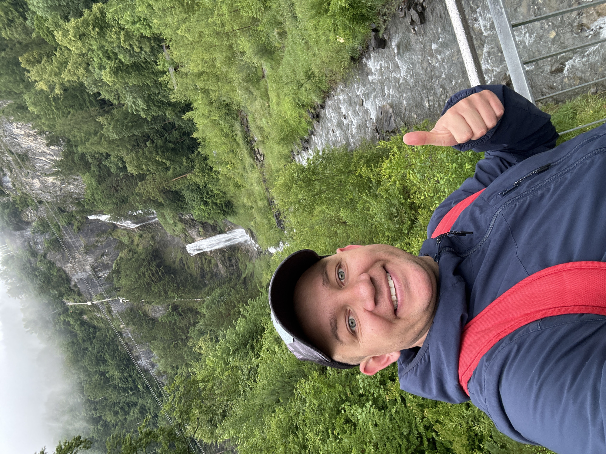 In a selfie taken outdoors, a smiling man with blue eyes and a baseball cap stands in front of a lush green natural landscape. He is wearing a dark blue hooded jacket with a red backpack over it, and he gives a thumbs-up gesture with his right hand. Behind him, a narrow, fast-flowing river runs through a rocky bed, flanked by dense green foliage. Further in the background, a tall, cascading waterfall plunges down a steep, rocky cliff face partially obscured by trees and mist. The overall scene suggests a scenic location, possibly in the Swiss Alps, given the mention of Hüsen, Switzerland. The environment is characterized by natural elements like water, rocks, trees, and a waterfall, with a slightly overcast or misty sky contributing to the atmospheric setting.