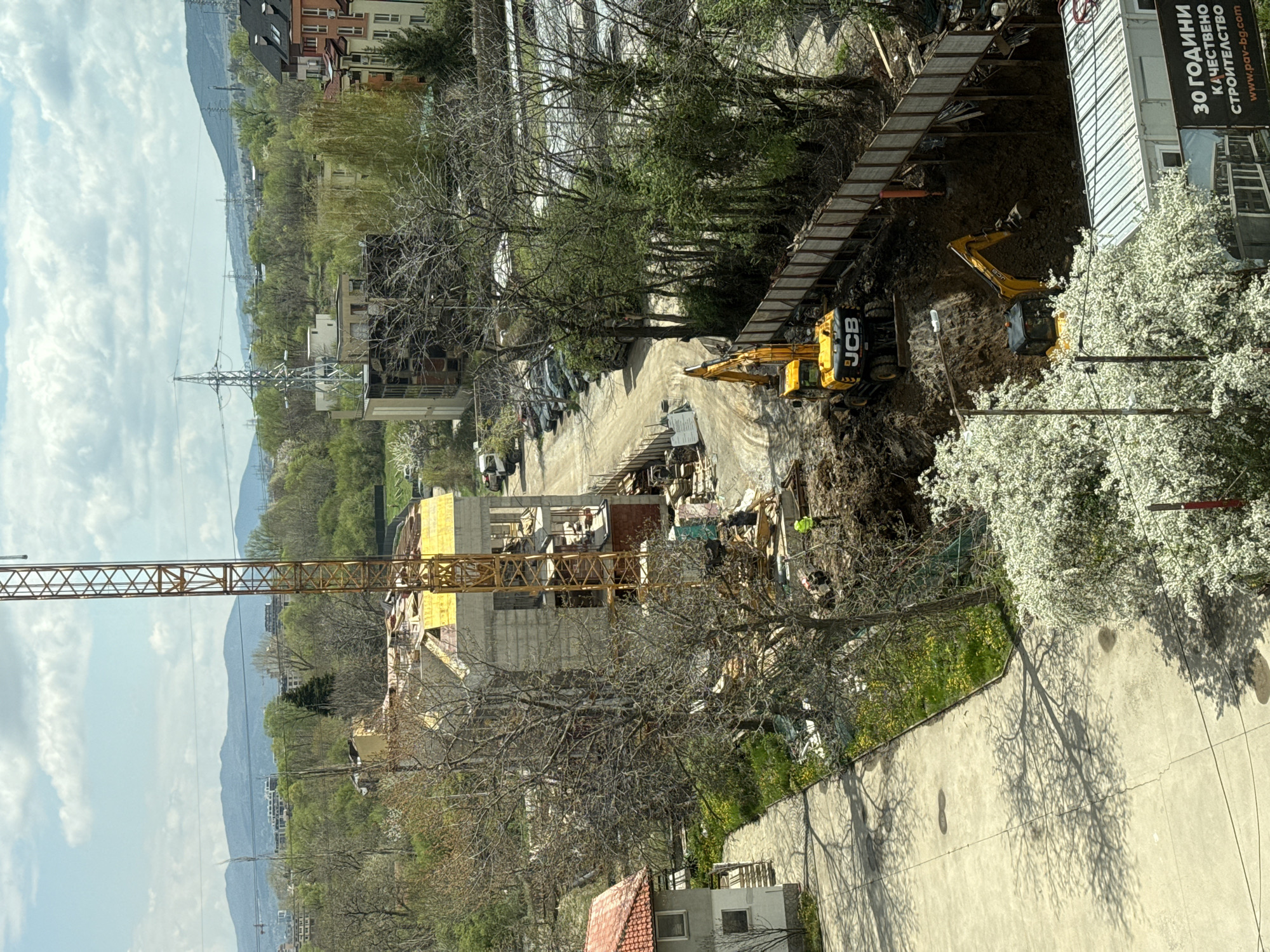 The image depicts a construction site in Sofia, Bulgaria. Two yellow JCB excavators are digging into the ground. A building under construction is visible with a crane to the left. There are trees, bushes and a flowering tree near the digging site. In the background, there are buildings, trees, a mountain, and power lines. A sign with white letters is visible to the right of the site.