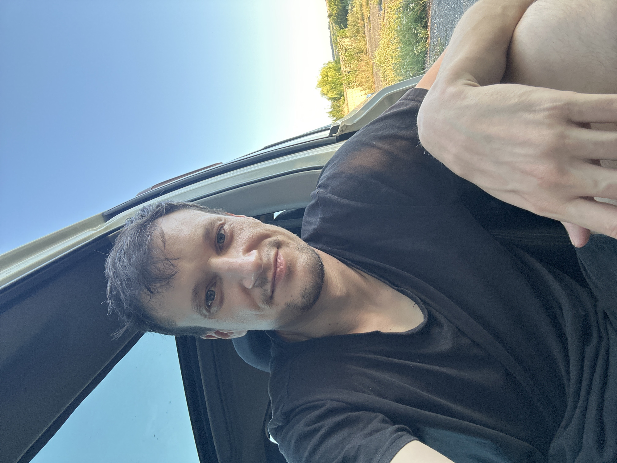 A medium shot selfie shows a man with short, dark hair and stubble sitting in the driver's seat of a vehicle with the sunroof open and the driver's side door ajar. He is wearing a black t-shirt and is looking towards the camera with a slight smile. His left arm is resting on his lap, and his right arm is bent at the elbow, with his hand resting on his leg. The background shows a bright blue sky and a rural landscape with some trees and vegetation. The image appears to have been taken during daylight hours. The metadata indicates the image was taken in Makiv, Ukraine.