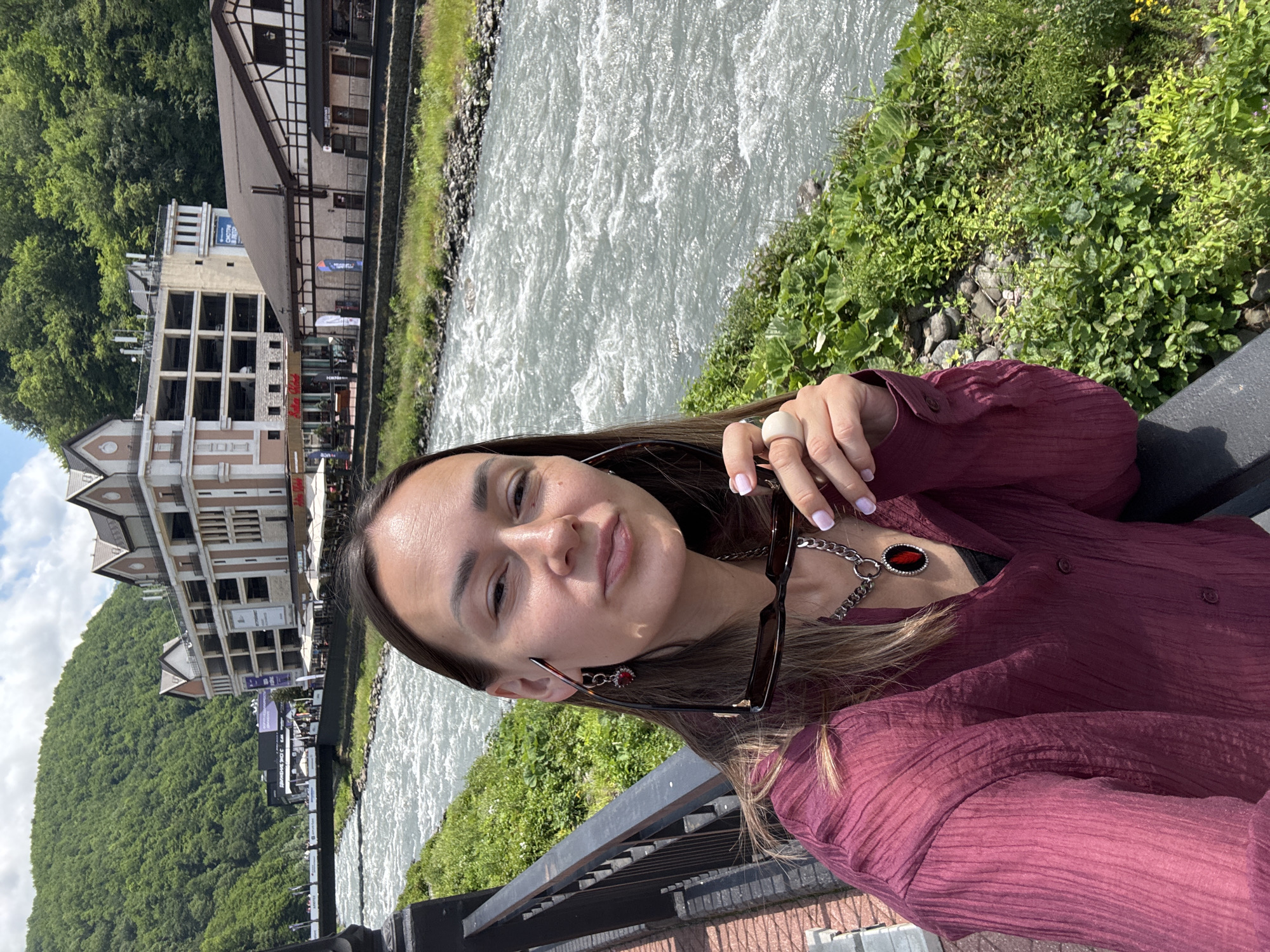 A woman with long brown hair is taking a selfie in front of a river and buildings in Estosadok, Russia. She is wearing a maroon collared shirt, a silver chain with a pendant, and a ring on her right hand. She is holding a pair of sunglasses in her right hand. The river is wide and appears to be flowing rapidly, with white water visible. In the background, there are several multi-story buildings and a lush green mountainside. The sky is partly cloudy. The woman is smiling and looking at the camera.