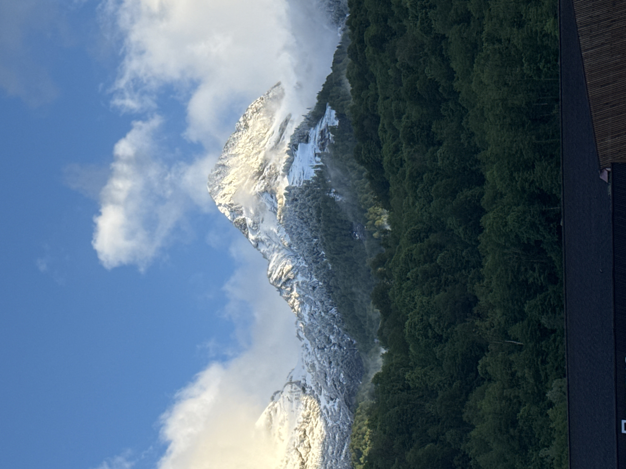 The image shows a snow-covered mountain peak adjacent to a dense, green forest under a blue sky with white clouds. Part of a dark building is visible on the right side of the image. The location is likely in a mountainous region, consistent with the topography around Estosadok, Russia.