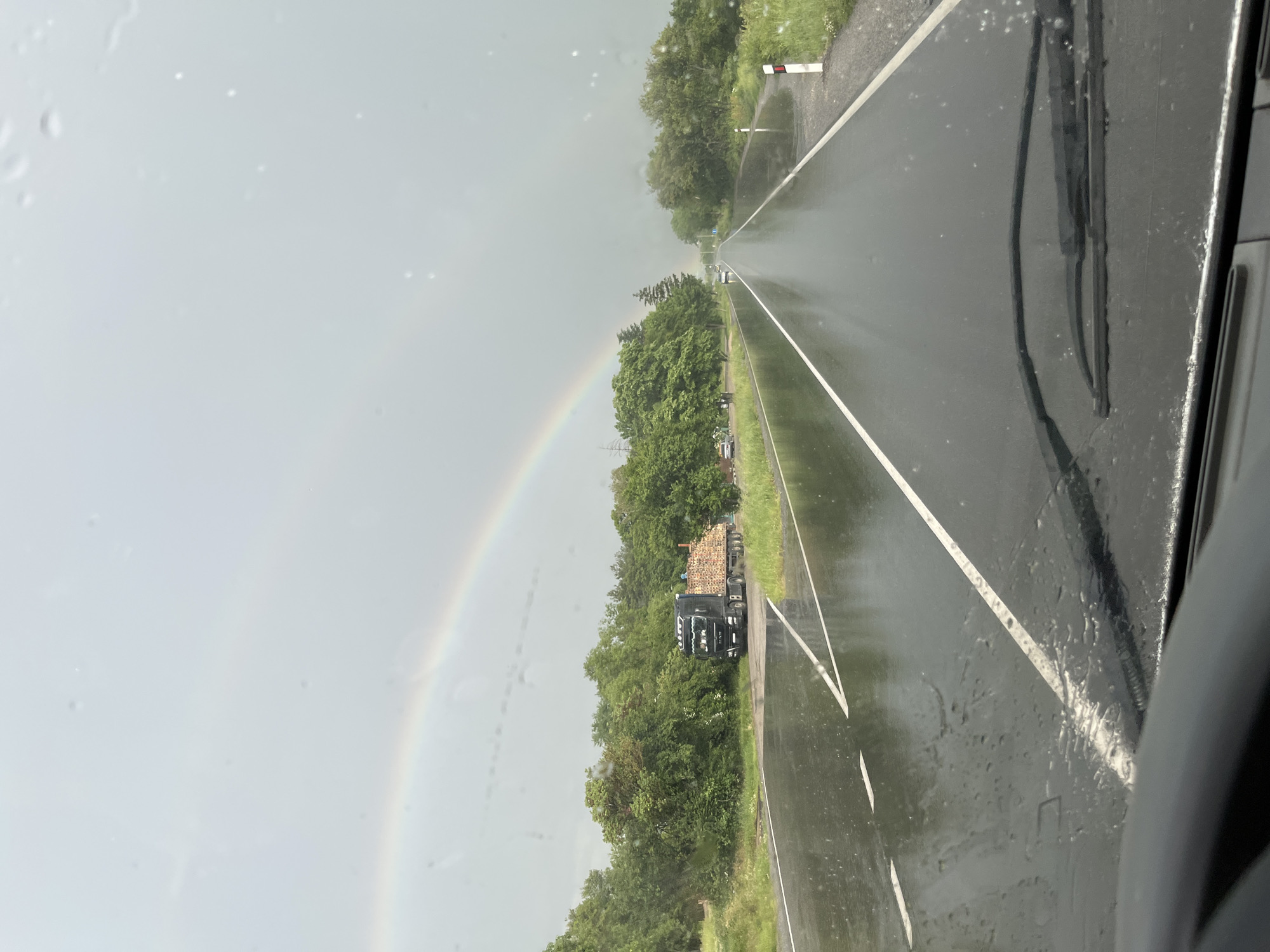 The image depicts a view from inside a vehicle driving on a wet road. A partial rainbow is visible in the sky to the left. On the right side of the road, a truck carrying logs is parked along the shoulder. There are trees along the side of the road. The location cannot be ascertained as Pevtsite, Bulgaria from the image alone. The scene is overcast and it is raining. Windshield wipers are visible on the vehicle. The image is rotated to display vertically.