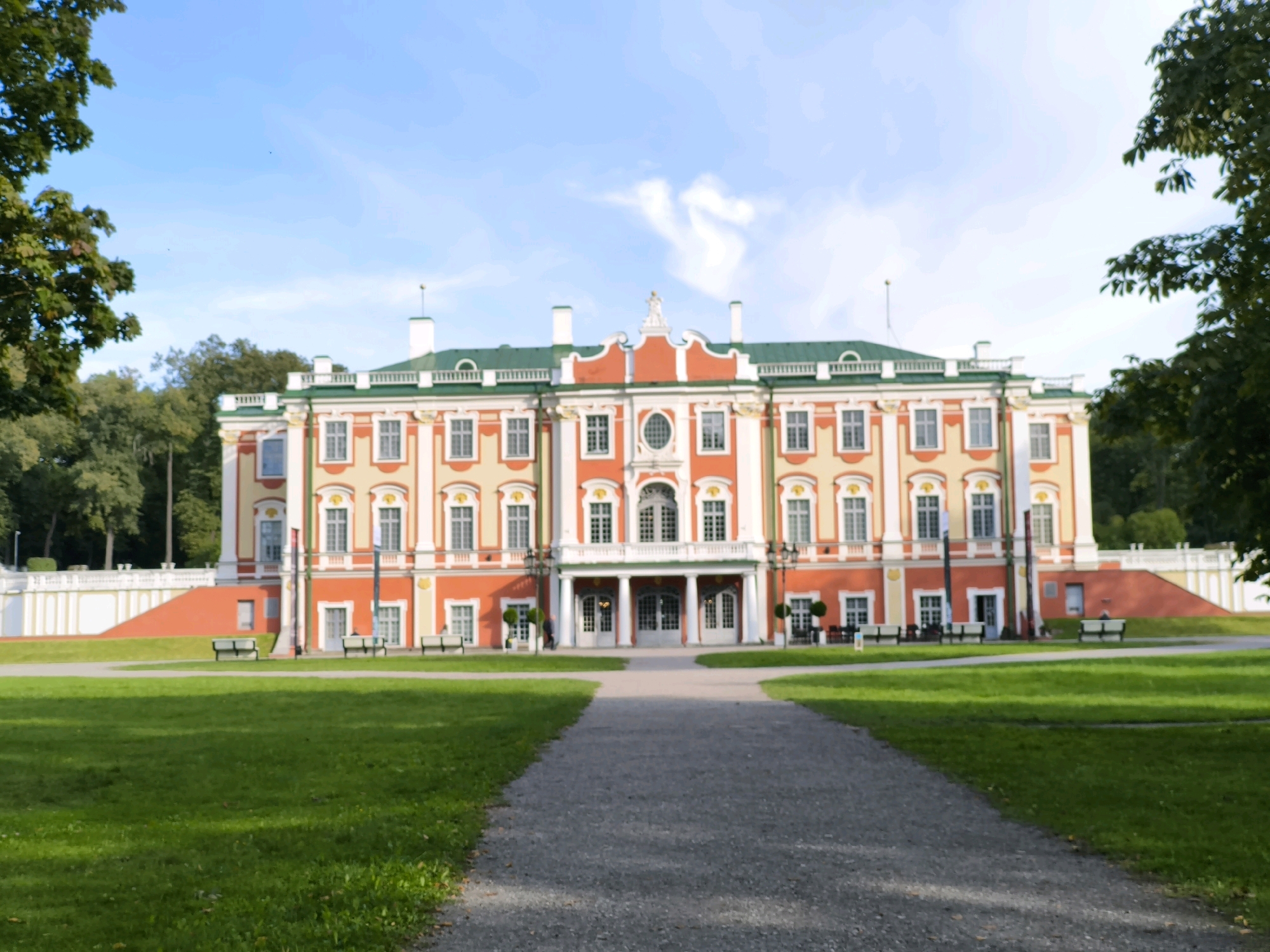 The image shows Kadriorg Palace, a Baroque palace in Tallinn, Estonia, situated in a park. The palace is a prominent building with a red, yellow, and green facade, featuring multiple windows and decorative elements. In front of the palace, a gravel path leads through a manicured green lawn. Several benches are visible on either side of the path. Trees frame the scene on the left and right sides, with a bright blue sky above. The overall impression is one of a well-maintained historical estate on a clear day. There are no people clearly visible in the image.