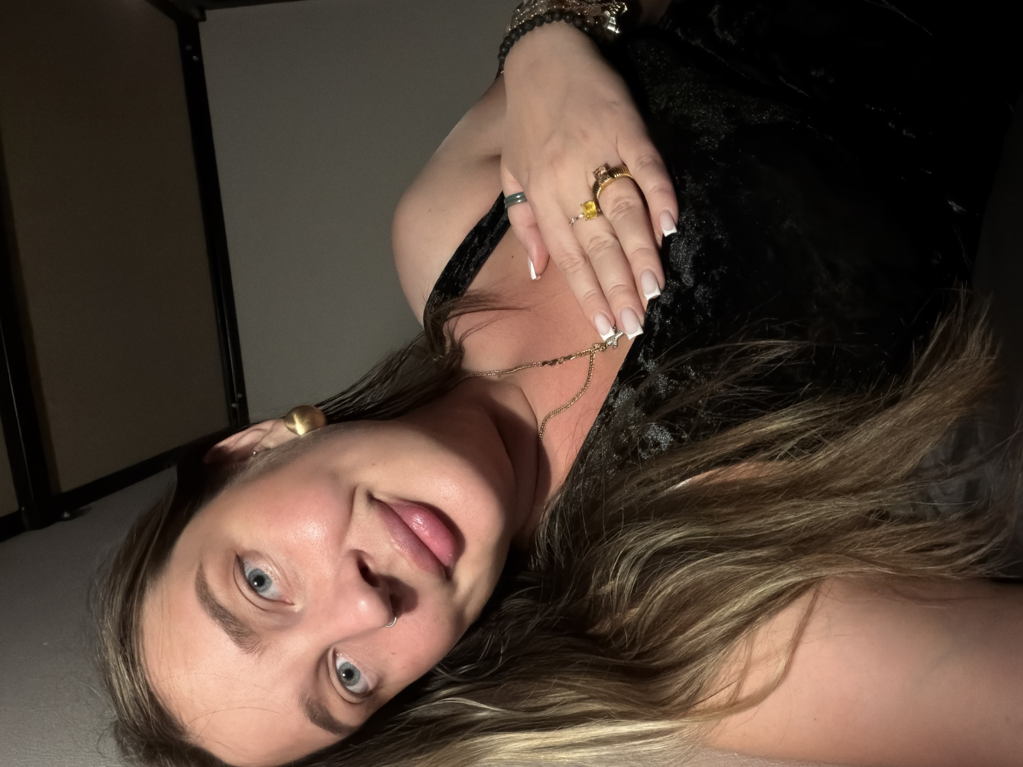 A woman with blonde hair, wearing a black top, is photographed from a low angle. She is wearing a gold necklace and multiple rings on her fingers. She has a nose piercing and a gold earring. The background is a dimly lit interior with a shelf in the corner. The location is not discernible.