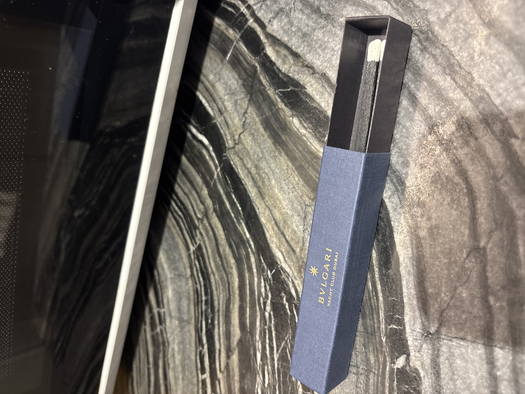 A navy blue box with gold lettering is placed on a dark, patterned countertop. The box is open, revealing a single match inside. The lettering on the box reads "BULGARI YACHT CLUB DUBAI". The background shows the edge of a dark appliance, likely a microwave, with a white trim. The countertop has a swirling, marbled pattern of black and grey. The image is a close-up, static shot taken from a slightly elevated angle. There are no people or any other discernible objects in the frame. The scene appears to be indoors.