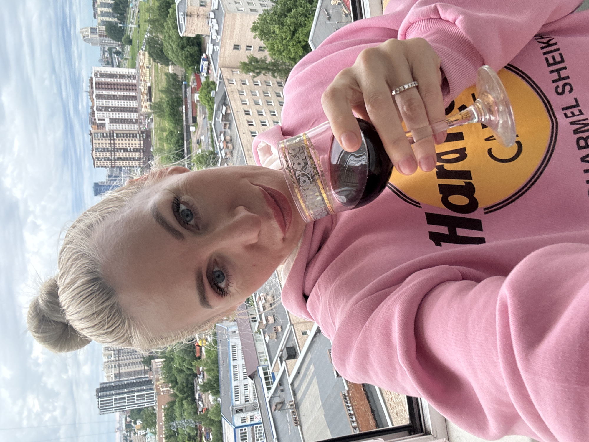 A blonde woman with her hair in a bun takes a selfie holding a glass of red wine. She is wearing a pink hoodie with a yellow circular logo that says "Hard Rock Cafe." The background shows a cityscape with multiple buildings and trees under a cloudy sky. She is wearing a ring on her left hand.