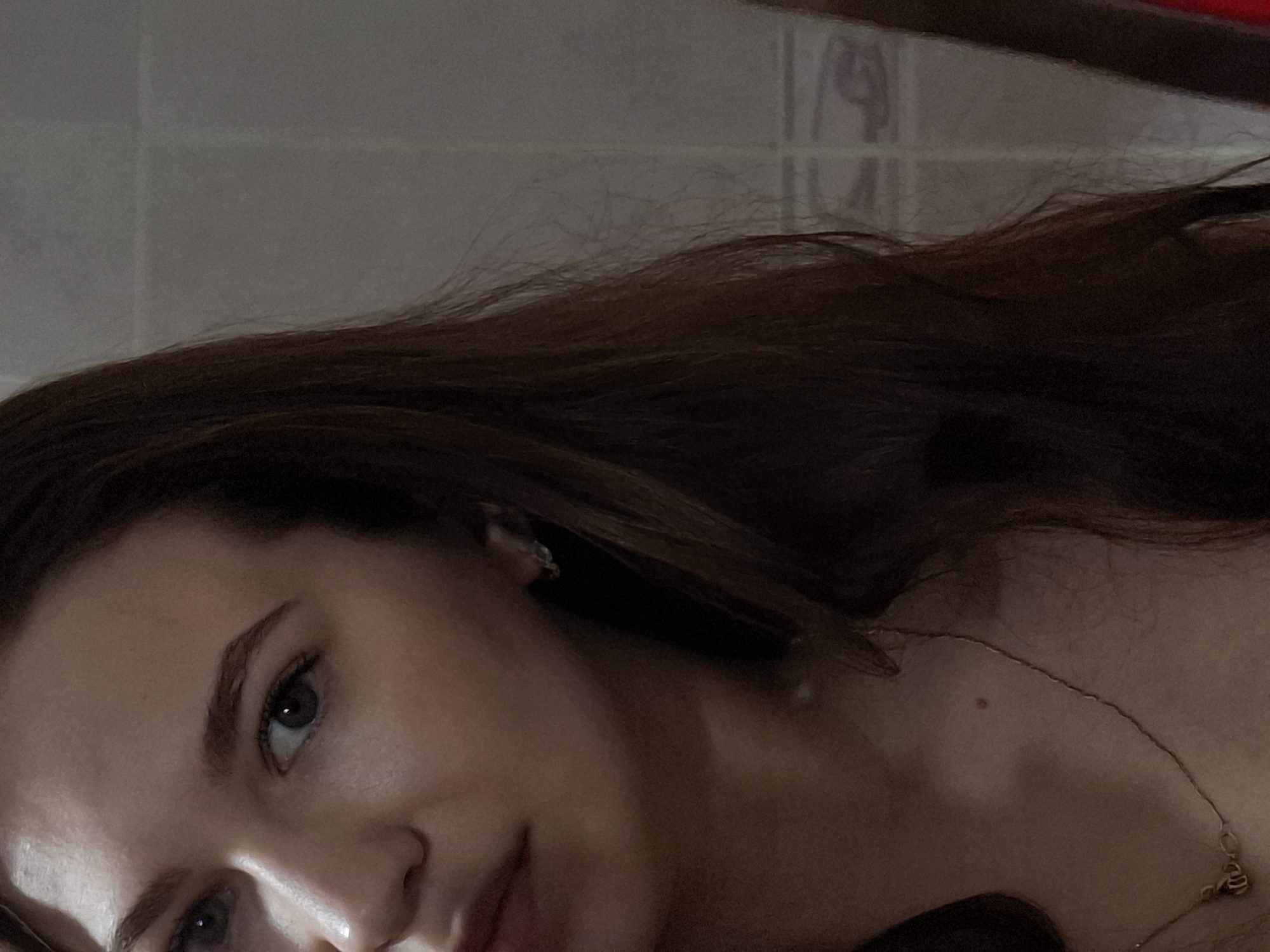 A close-up shot of a woman with long brown hair, facing the camera at an angle. She has fair skin and green eyes. A delicate gold necklace is visible around her neck. The background appears to be a light-colored tiled wall. There's no discernible information in the image to indicate it was taken in Chaikovsky, Russia.