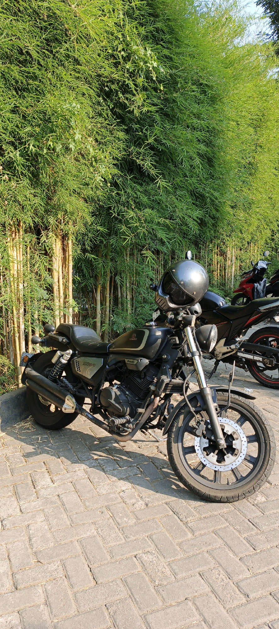 A black motorcycle with a helmet on its handlebars is parked on a brick pathway in front of a dense bamboo forest. The motorcycle is the primary subject of the image, with its front wheel, engine, seat, and exhaust pipe clearly visible. A second, red motorcycle is partially visible behind the black one. The bamboo forest creates a lush green backdrop. The pathway is made of interlocking rectangular bricks arranged in a herringbone pattern, with some dry leaves scattered on the ground. The lighting suggests it is daytime, with sunlight casting shadows on the pathway. The scene appears to be an outdoor setting in Trihanggo, Indonesia, likely a park or a roadside area with natural vegetation.