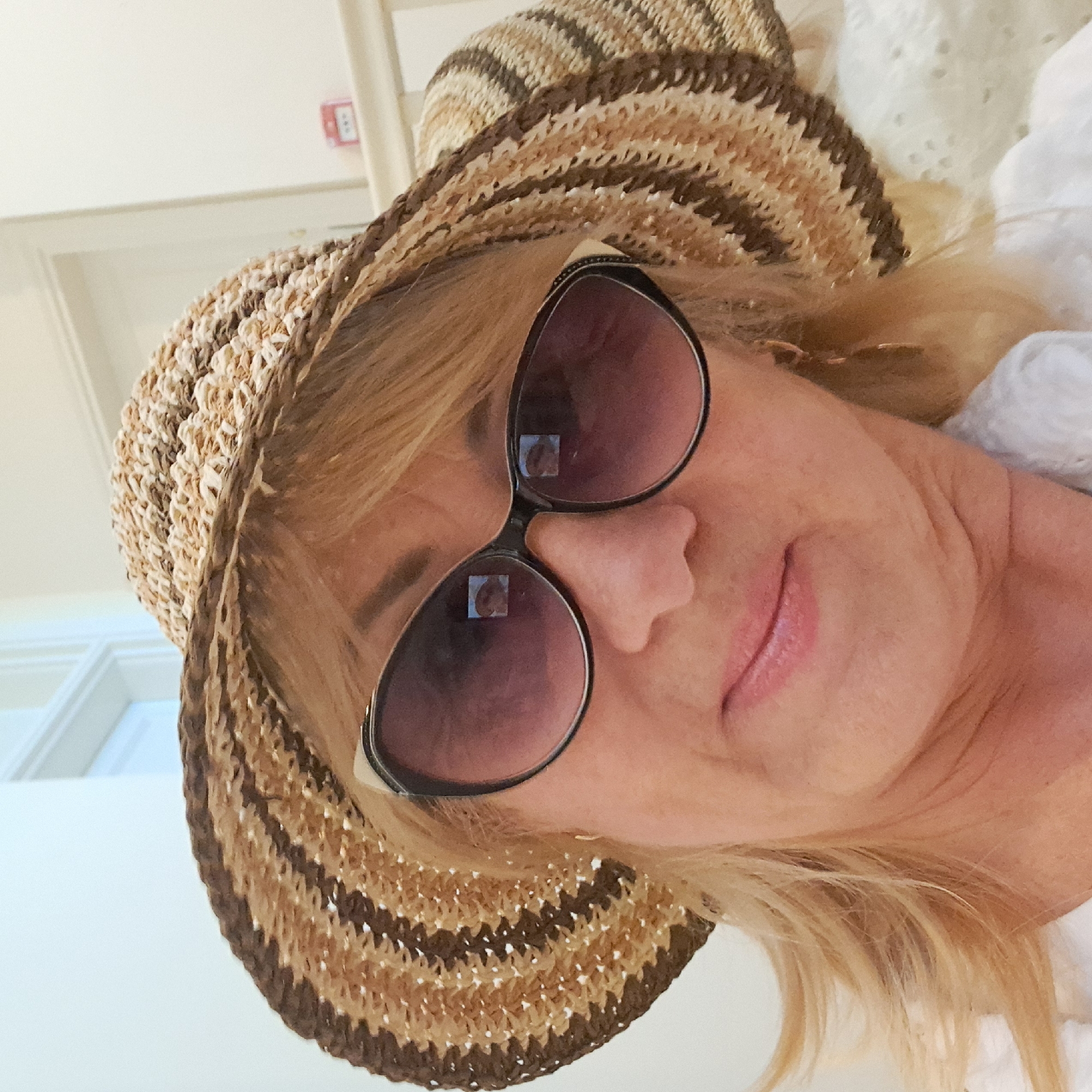 This media file shows a close-up, rotated portrait of a blonde woman wearing a straw hat and sunglasses. She is looking towards the camera with a slight smile. The sunglasses have a dark, reflective lens, and the frames are black and white. Her hair is blonde and appears to be layered. She is wearing a white top, possibly made of a light, breathable fabric with eyelet detailing. The background is out of focus, but appears to be an indoor setting with white walls and what looks like a doorway or window frame on the left. There is a small red fire alarm on the wall in the upper left corner. The lighting suggests it is daytime. The overall mood is relaxed and casual. The media file does not contain any visible text, apart from a small, blurry reflection of a person in the sunglasses.