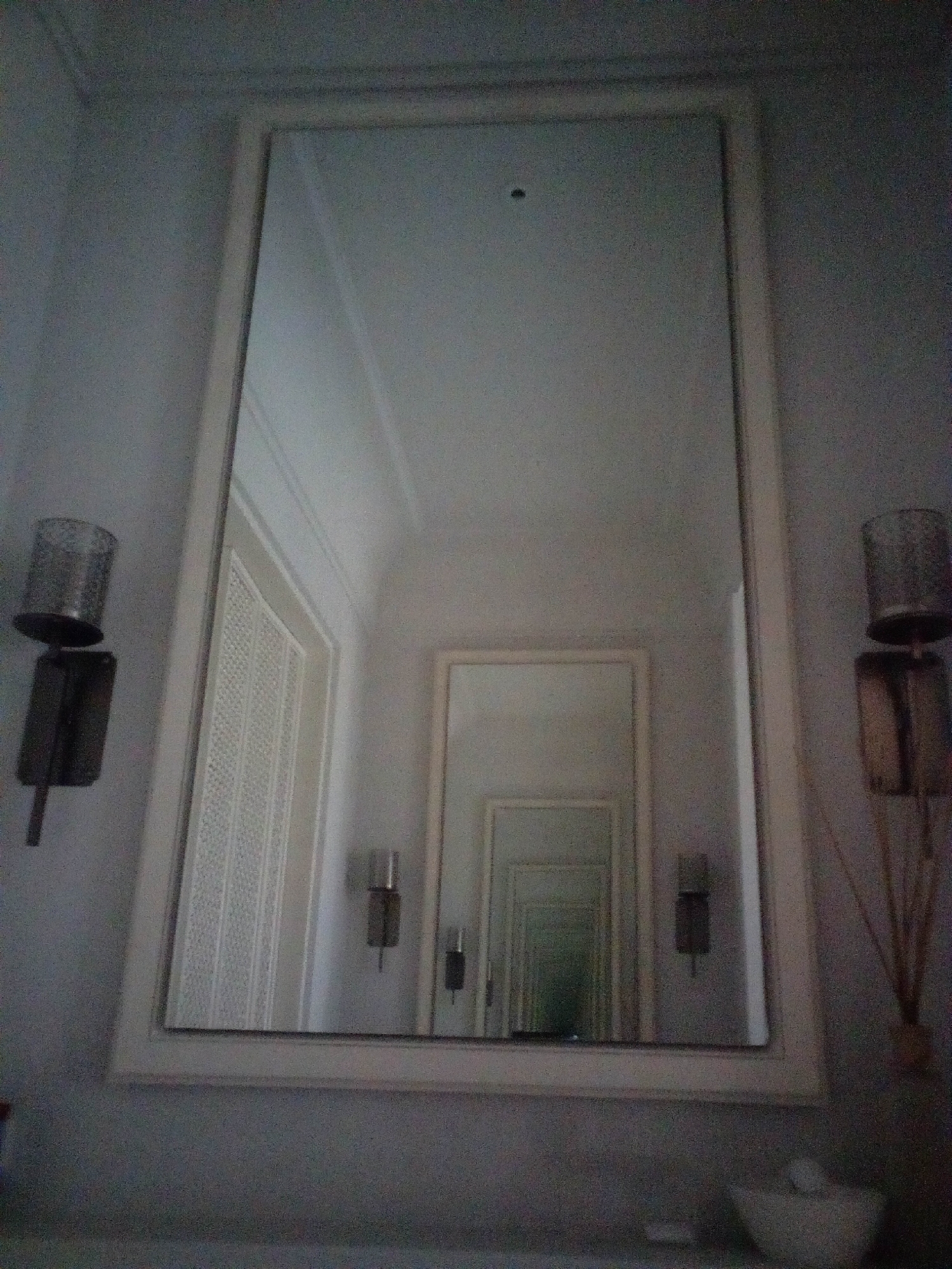 The image shows a large, ornate mirror on a wall, reflecting a hallway with multiple doors. On the left side of the mirror, there is a sconce light fixture with a cylindrical shade. On the right side of the mirror, another sconce light fixture is visible, along with what appears to be a diffuser with reeds. The mirror's reflection creates a sense of depth, showing a repeating pattern of doorways. The overall lighting is dim, suggesting an indoor setting. The specific location in South Jakarta, Indonesia, is not discernible from the visual information provided in the image.