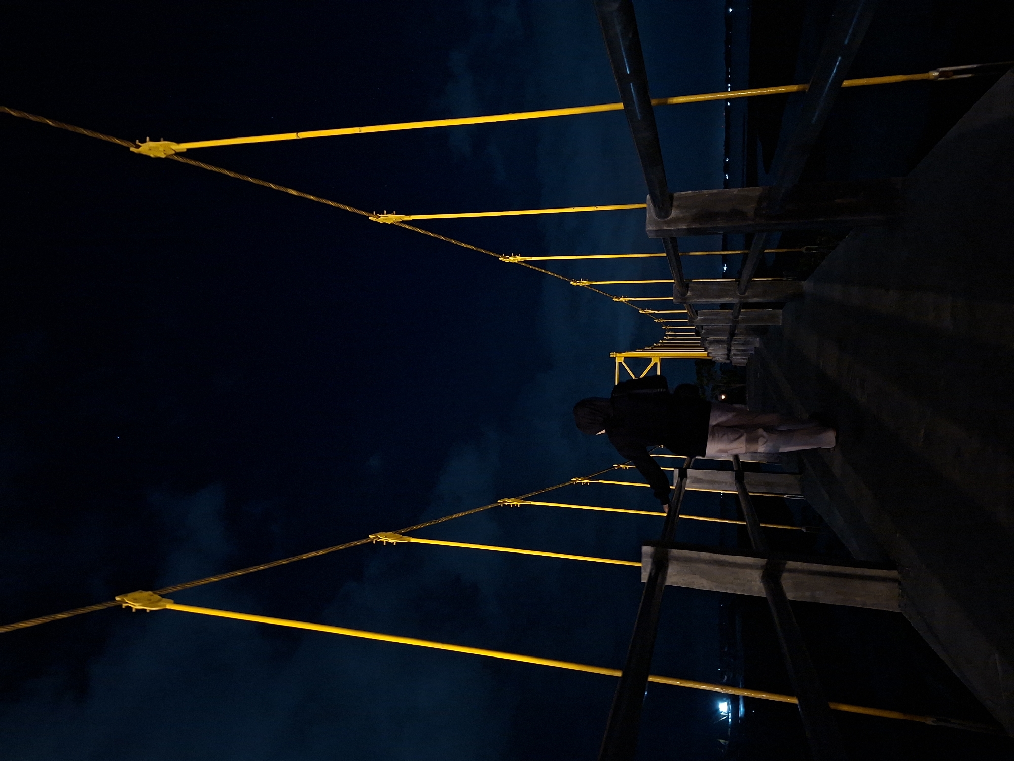 A person in dark clothing and light-colored pants stands on the side of a pedestrian bridge at night in Lhokseumawe, Indonesia. The bridge features yellow support structures and metal railings. The dark sky is visible in the background.