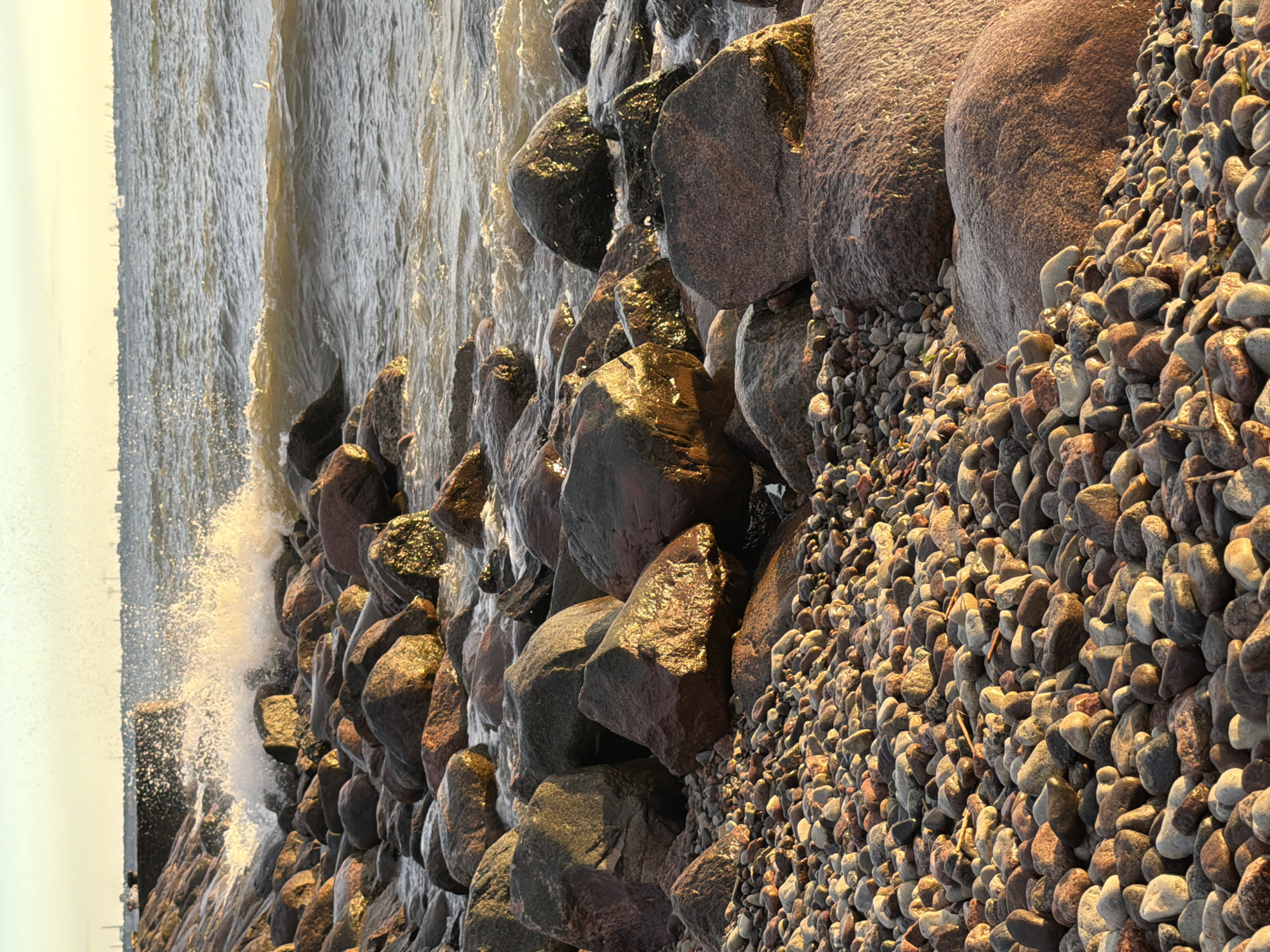 The image depicts a rocky shoreline with waves crashing against large boulders and smaller pebbles. The water is choppy and reflects the warm, golden light of what appears to be either sunrise or sunset, casting a glow on the wet rocks. In the background, a hazy line of trees or buildings suggests a distant landmass, with a few indistinct figures visible near the water's edge. The overall scene conveys a sense of natural beauty and the dynamic power of the sea. The location is identified as Tallinn, Estonia. No text is visible in the image.