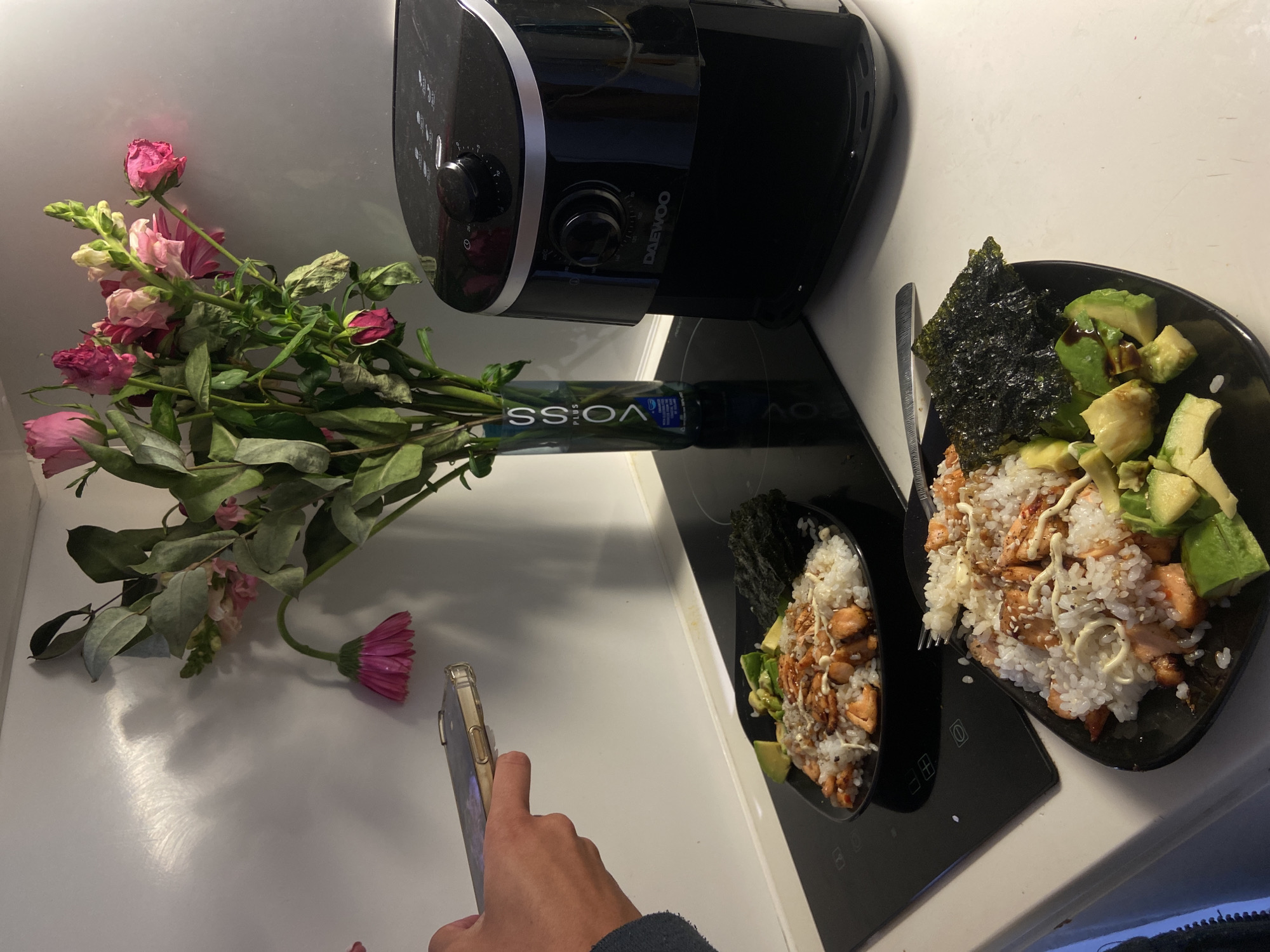 A countertop scene features a black Daewoo air fryer, a clear glass vase with pink and green flowers, and two black bowls of food on a black stovetop. The food appears to be a rice dish with avocado, seaweed, and some type of protein drizzled with a white sauce. A hand holding a smartphone is visible in the lower left, suggesting someone is taking a photo. The setting seems to be indoors, possibly a kitchen. The location within London, United Kingdom, cannot be determined from the image.