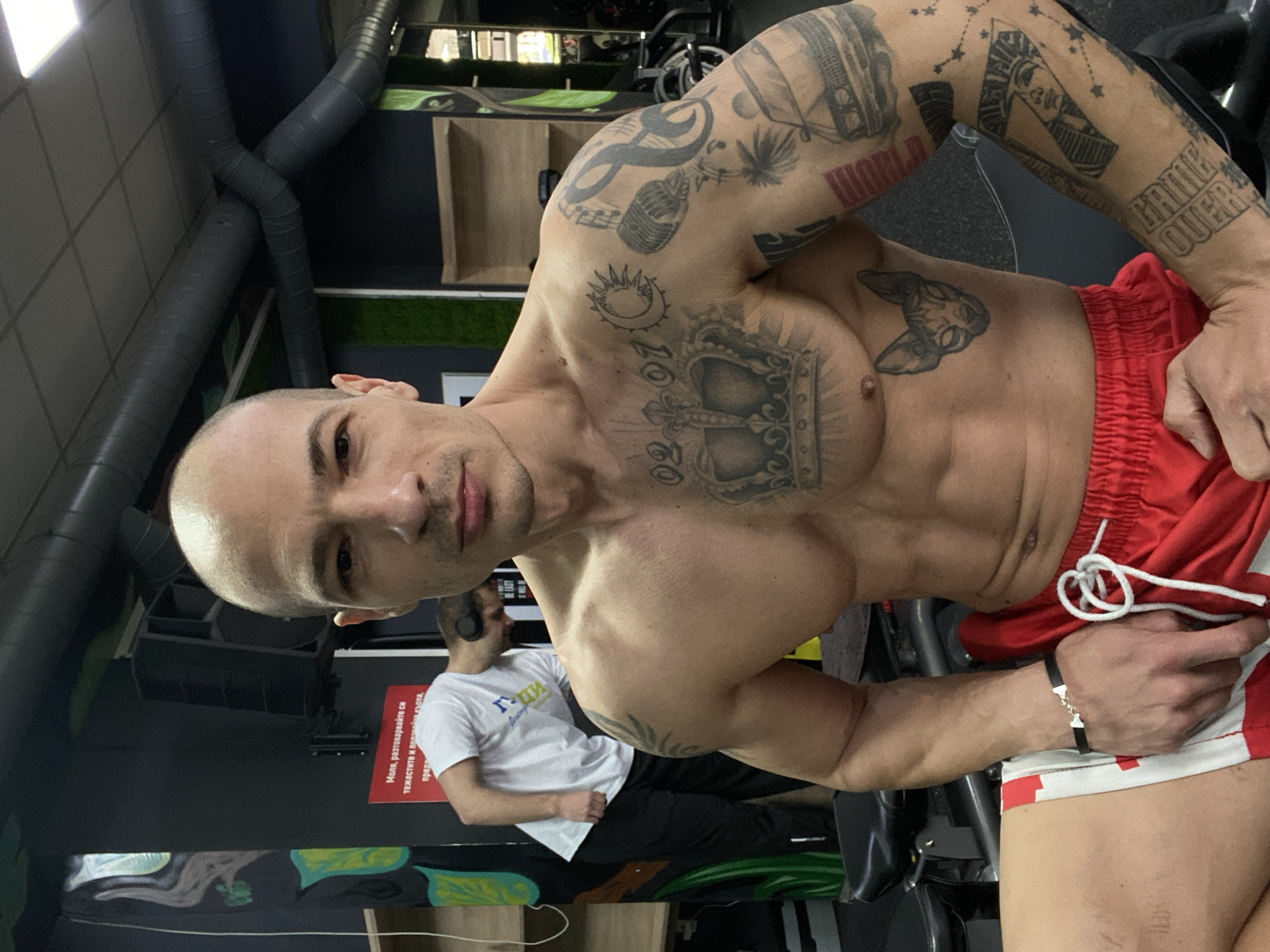 A shirtless man with numerous tattoos on his arms and chest is posing in a gym. He is wearing red and white shorts and a black bracelet. Another man in a white t-shirt and black pants is partially visible in the background. The location appears to be a gym, evidenced by the exercise equipment, pipes on the ceiling, and what may be part of a weight rack. There's a sign on the wall in Bulgarian, which may indicate a location in Pleven, Bulgaria.