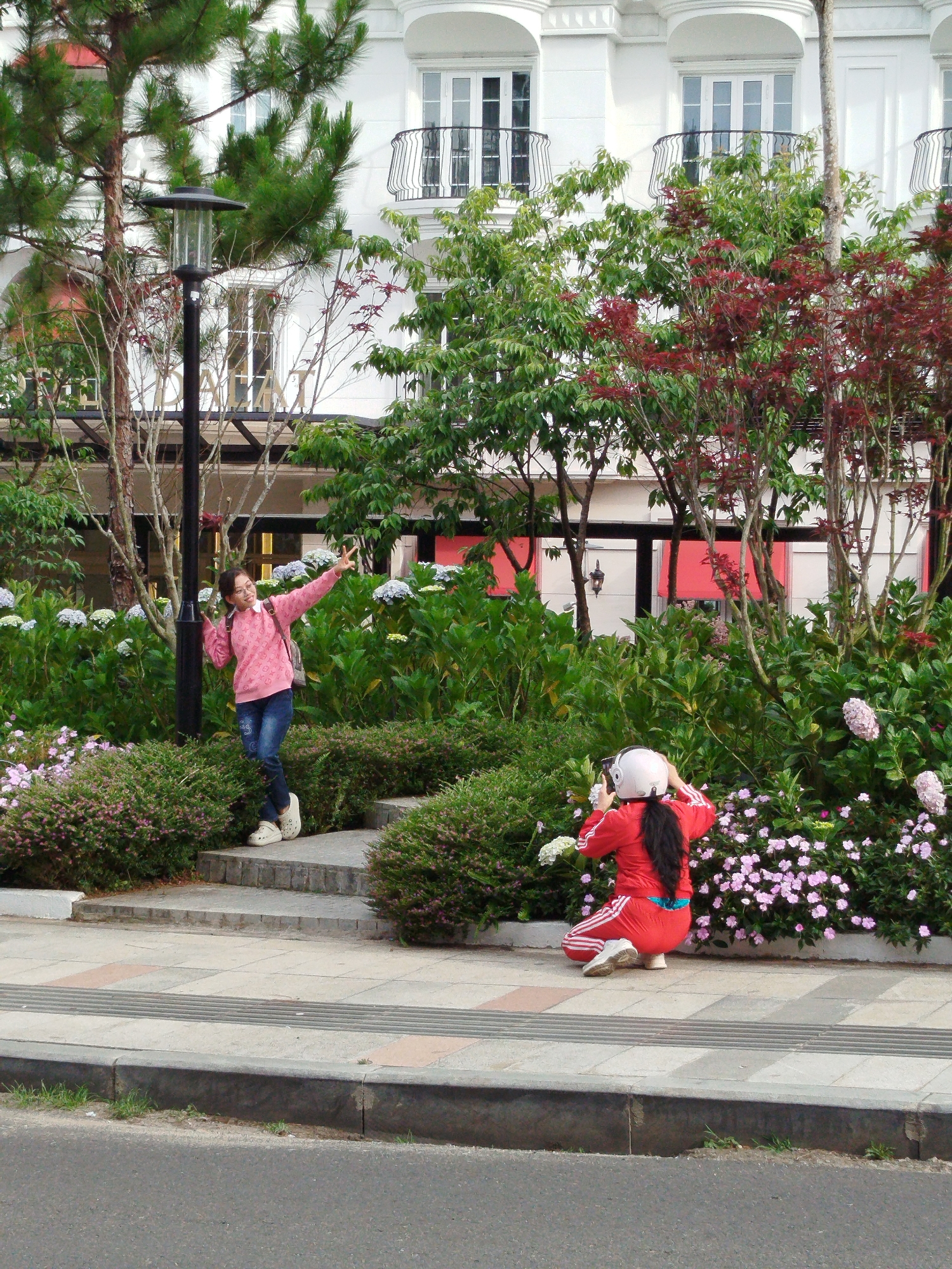 A full outdoor shot shows two people and a background of trees and buildings. A light-skinned person wearing a pink long-sleeved shirt and jeans stands on a set of concrete stairs, with one hand extended upward in a "V" sign. They have a backpack and wear white shoes. A second person, facing away from the camera, is crouched down on the sidewalk, wearing a red tracksuit and a white helmet. They appear to be taking a photo with a camera.
The background consists of lush greenery with various shrubs and flowers, a black lamppost, and a multi-story white building with balconies and window boxes. A red awning is visible behind the greenery. The location is likely Da Lat, Vietnam, based on the architecture, plant life, and general environment. The photo seems to capture the act of taking a photograph in a tourist setting.