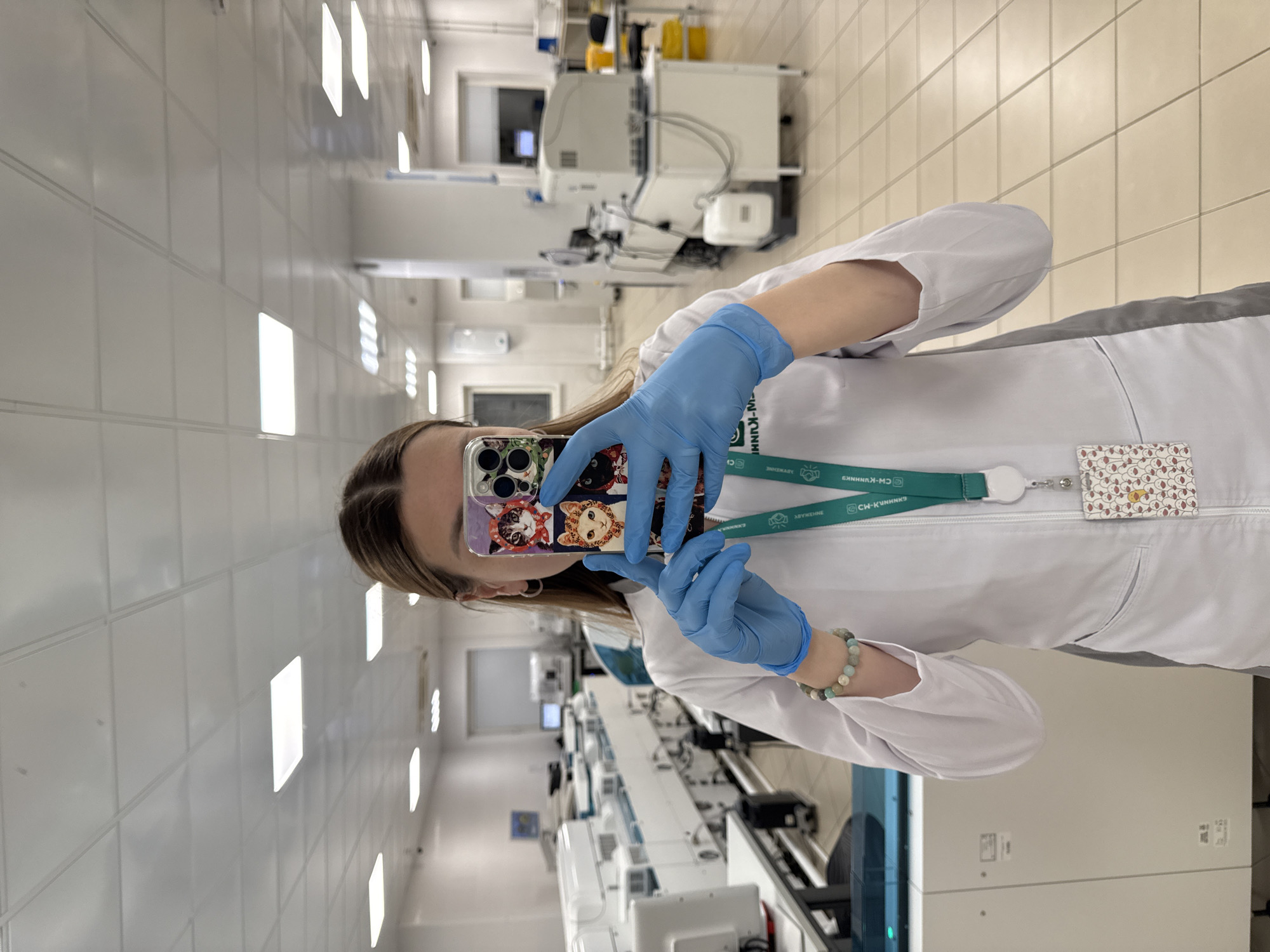 A person, likely female, is depicted taking a selfie indoors within a modern laboratory or medical facility. The image is presented rotated 90 degrees clockwise. The individual wears a white lab coat, light blue nitrile gloves, and a green lanyard around their neck. A beaded bracelet adorns their left wrist. They hold a smartphone with a distinctive case featuring colorful cartoon cat-like characters.
The background shows a brightly lit, sterile environment with white tiled walls and beige tiled floors. Various pieces of advanced laboratory equipment, including automated systems, processing machines, and computer monitors, are arranged along workbenches. The general appearance of the space suggests a high-tech medical or research setting, characteristic of a clinic or diagnostic center.
Visible text on the green lanyard includes "СИ-КЛИНКИ" (likely "CI-CLINICS") in Cyrillic, "CLINICS" in English, and other smaller Cyrillic words such as "НОВЫЕ" (meaning "NEW"), along with associated logos. A small, patterned card holder is clipped to the lanyard. The Cyrillic text, in conjunction with the provided context, indicates the location is in Moscow, Russia. The individual appears to be engaging in a moment of personal photography within their professional setting.