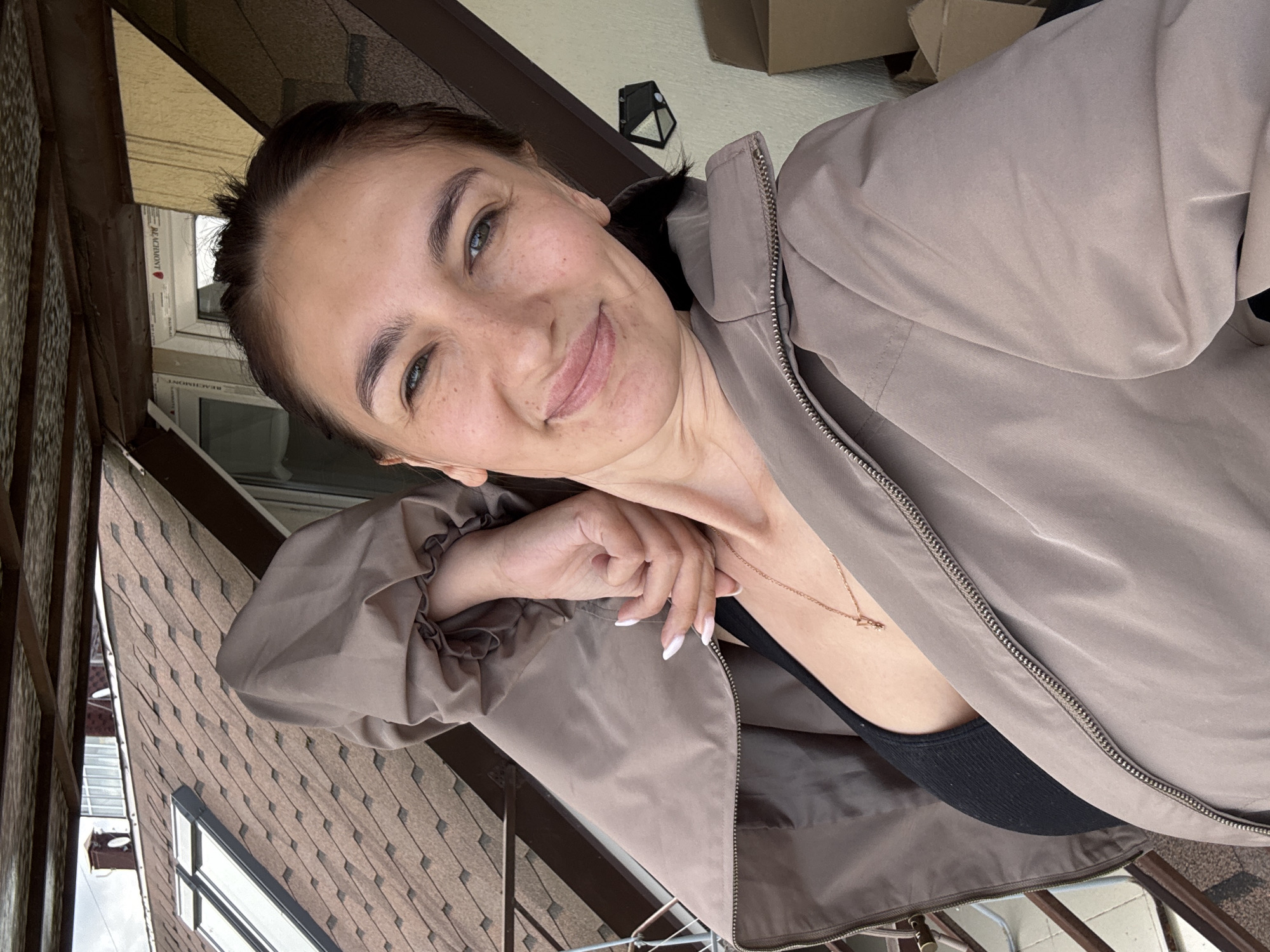 A woman is posing for a selfie outdoors in Sochi, Russia. She is wearing a taupe-colored jacket, unzipped to reveal a black top underneath. A gold necklace with a pendant is visible around her neck. Her hair is pulled back, and she has light skin with freckles. She is smiling slightly with a relaxed expression.
In the background, the environment suggests an urban setting. A building with brown shingled roofing and windows can be seen, and portions of other buildings are visible. There's a glimpse of a white window frame with the word "REACHMONT" on it. Above, there is a brown overhang and a box. The overall impression is that she is on a balcony or elevated platform. The image is rotated slightly, adding to the casual, candid feel of the photograph.