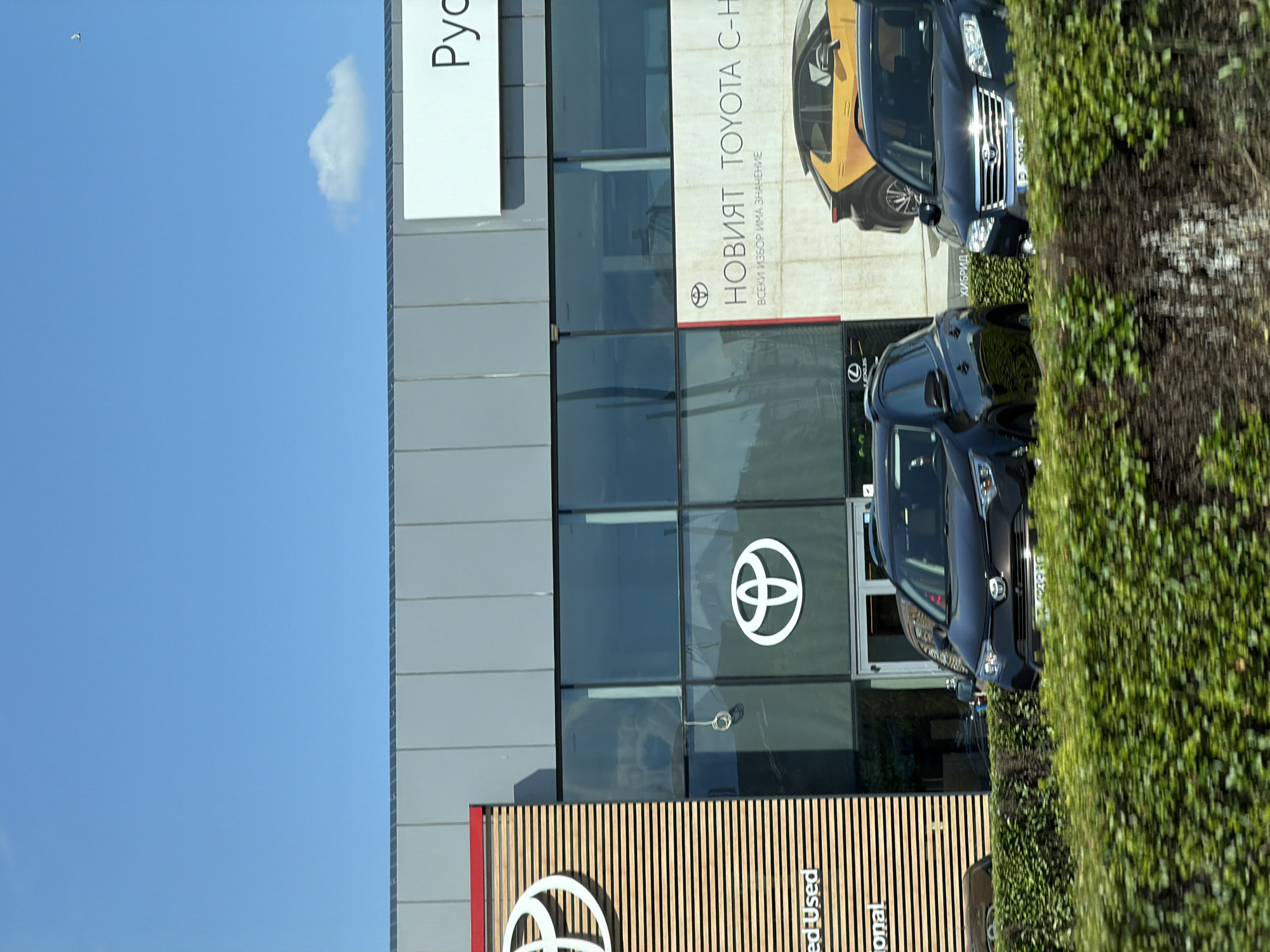 The image shows a Toyota dealership in Dolno Ablanovo, Bulgaria. The dealership has a large glass facade with the Toyota logo visible on the exterior. Inside, several cars are on display. In front of the dealership, there are several cars parked on the street, including a black Toyota RAV4 and a dark-colored SUV. The dealership is surrounded by a green hedge. The sky is blue with a few clouds. The overall impression is that of a well-maintained and modern car dealership.