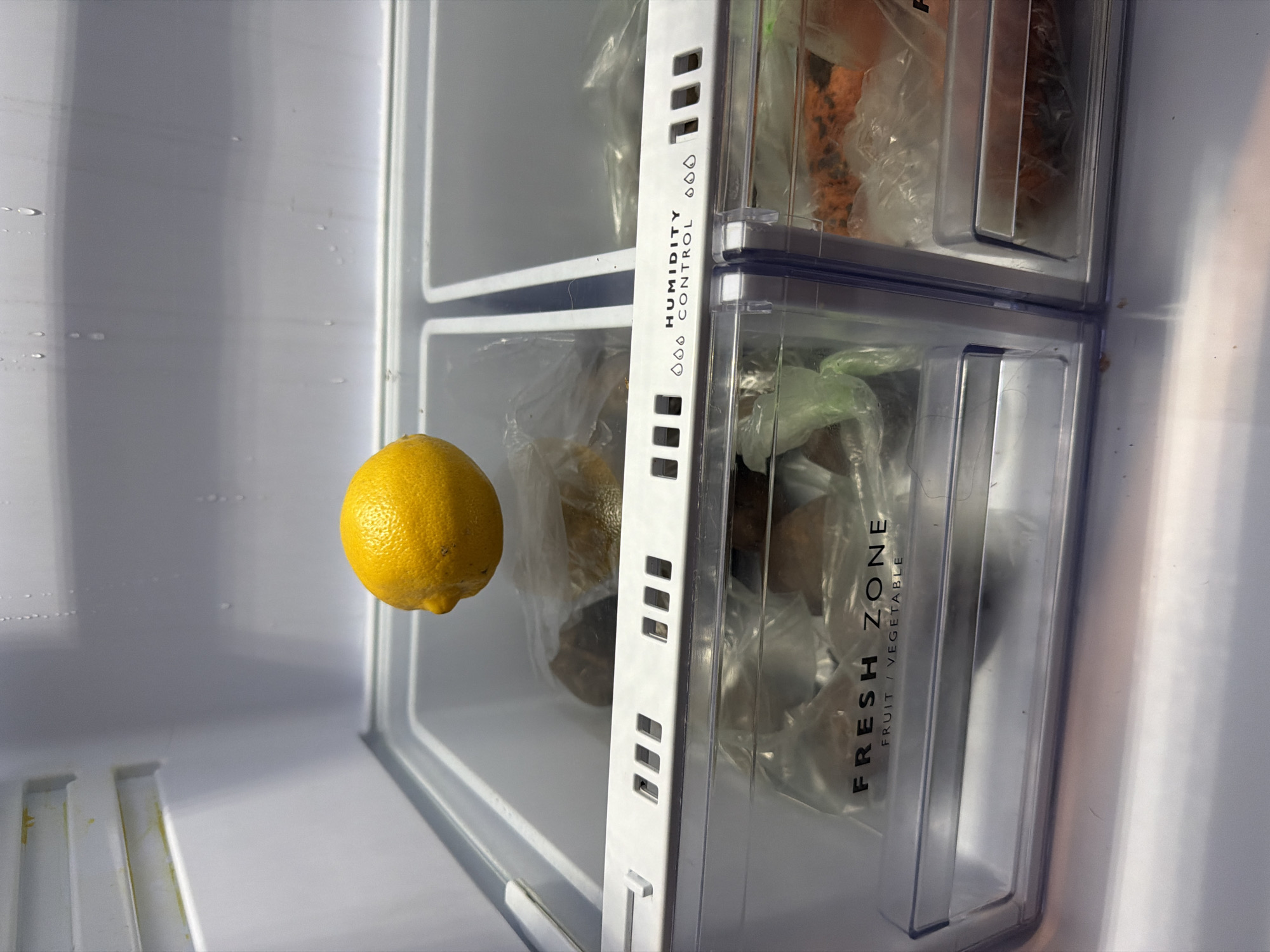 An overhead view captures the interior of a refrigerator compartment. A single, bright yellow lemon is suspended in mid-air, positioned centrally within the frame, above two plastic-lined drawers. The drawers are labeled "FRESH ZONE FRUIT / VEGETABLE" and "HUMIDITY CONTROL". The left drawer contains produce wrapped in plastic, and the right drawer also holds items in plastic wrapping, including what appears to be shredded carrots or similar vegetables. The refrigerator's interior walls are white and show signs of condensation. The overall environment is clean and organized, typical of a food storage appliance. The location in Sirius, Russia, is irrelevant to the visual content of the image.