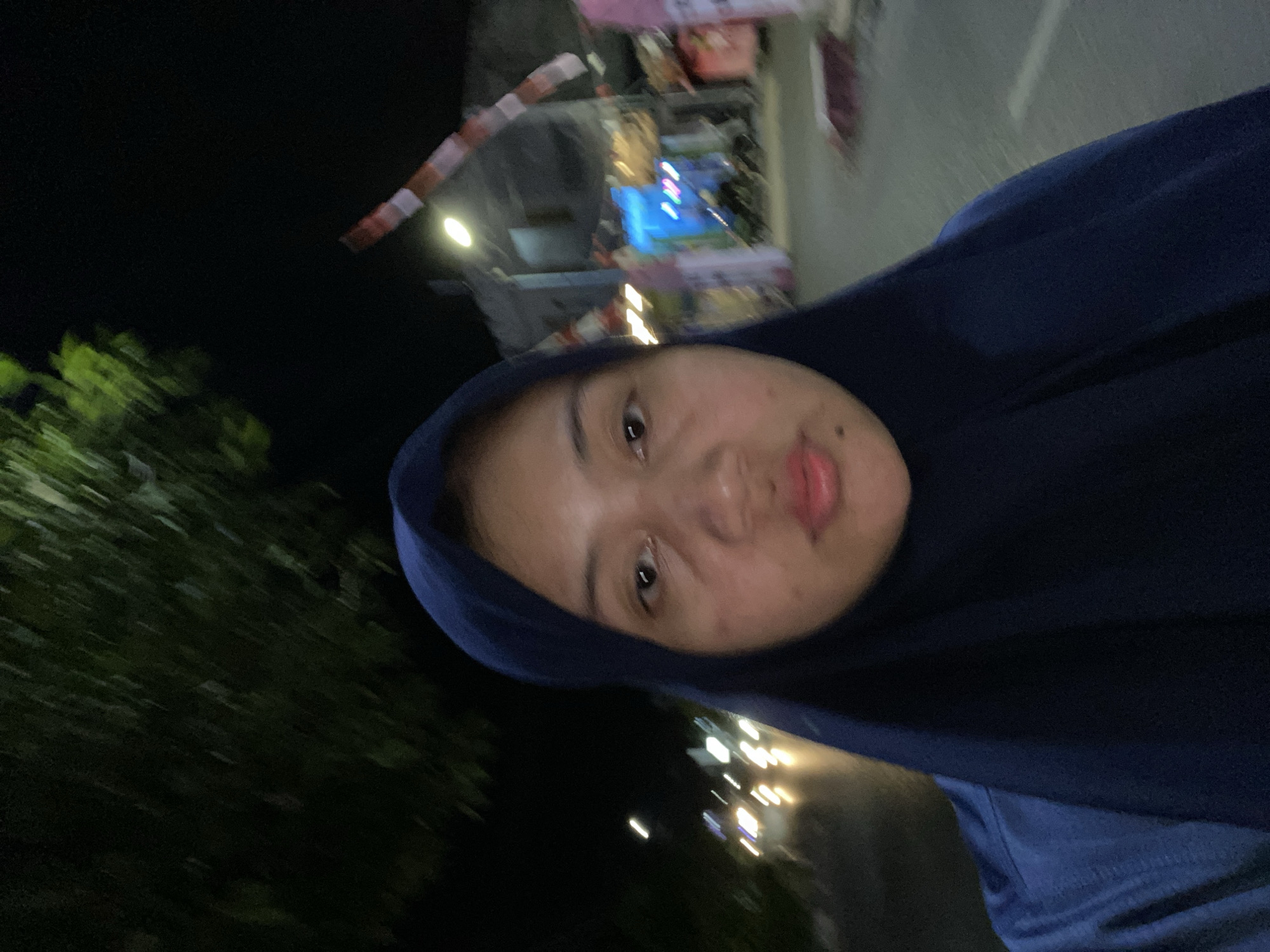 A slightly out-of-focus, selfie-style medium shot captures a young woman wearing a dark blue hijab. She is outdoors at night, with blurred lights of passing vehicles in the background, suggesting a street scene in the city of Banjarbaru, Indonesia. The background also shows hints of buildings and foliage, illuminated by streetlights and neon signs, one of which displays blue light. The woman's face is the primary focus, with her eyes open and looking towards the camera, and her lips slightly parted. Her skin appears to have some texture and discoloration. A red and white striped object, possibly a barrier or decoration, is visible further in the background.