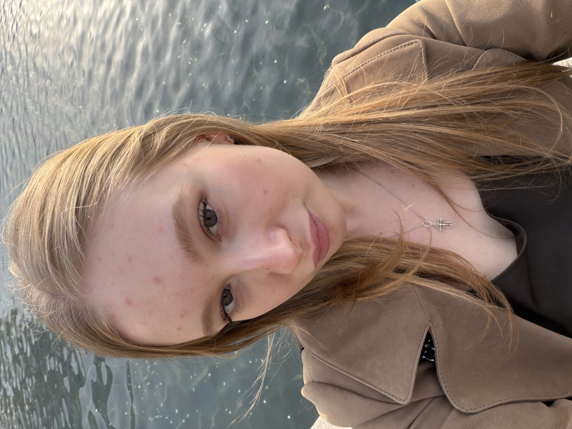 A young, light-skinned female with long, light brown hair is centered in the frame, slightly angled. She is wearing a tan jacket, and a silver necklace with a cross pendant is visible. Her gaze is directed at the camera. The background is a body of water, possibly a river or lake, with sunlight glinting off the surface. The location cannot be confidently determined as Krasnodar, Russia without further context or identifiable landmarks. No actions or interactions are depicted. The scene is outdoors and appears to be taken during the daytime.