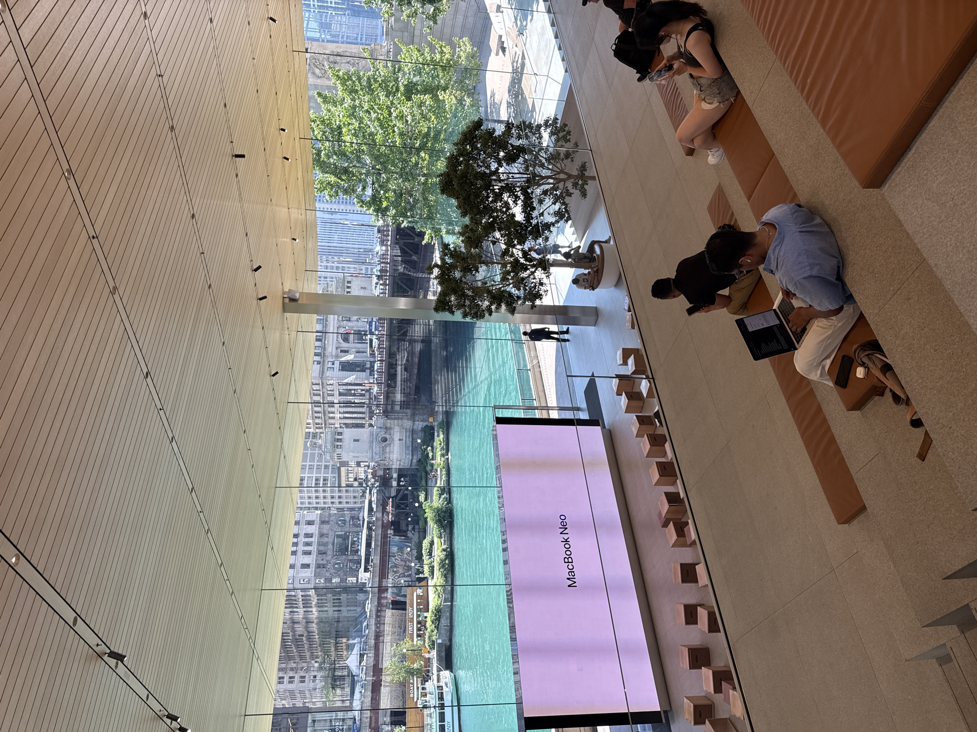 The image shows the interior of an Apple Store in Chicago, USA, with a view of the Chicago River through a large glass window. In the foreground, there are tiered seating areas with people using laptops and phones. A large screen displays "MacBook Neo." Outside, the Chicago River is visible with boats and buildings lining its banks, including the historic Michigan Avenue Bridge. Trees and other greenery are also present outside the store. The store's interior features a modern, minimalist design with wooden seating and a slatted ceiling.