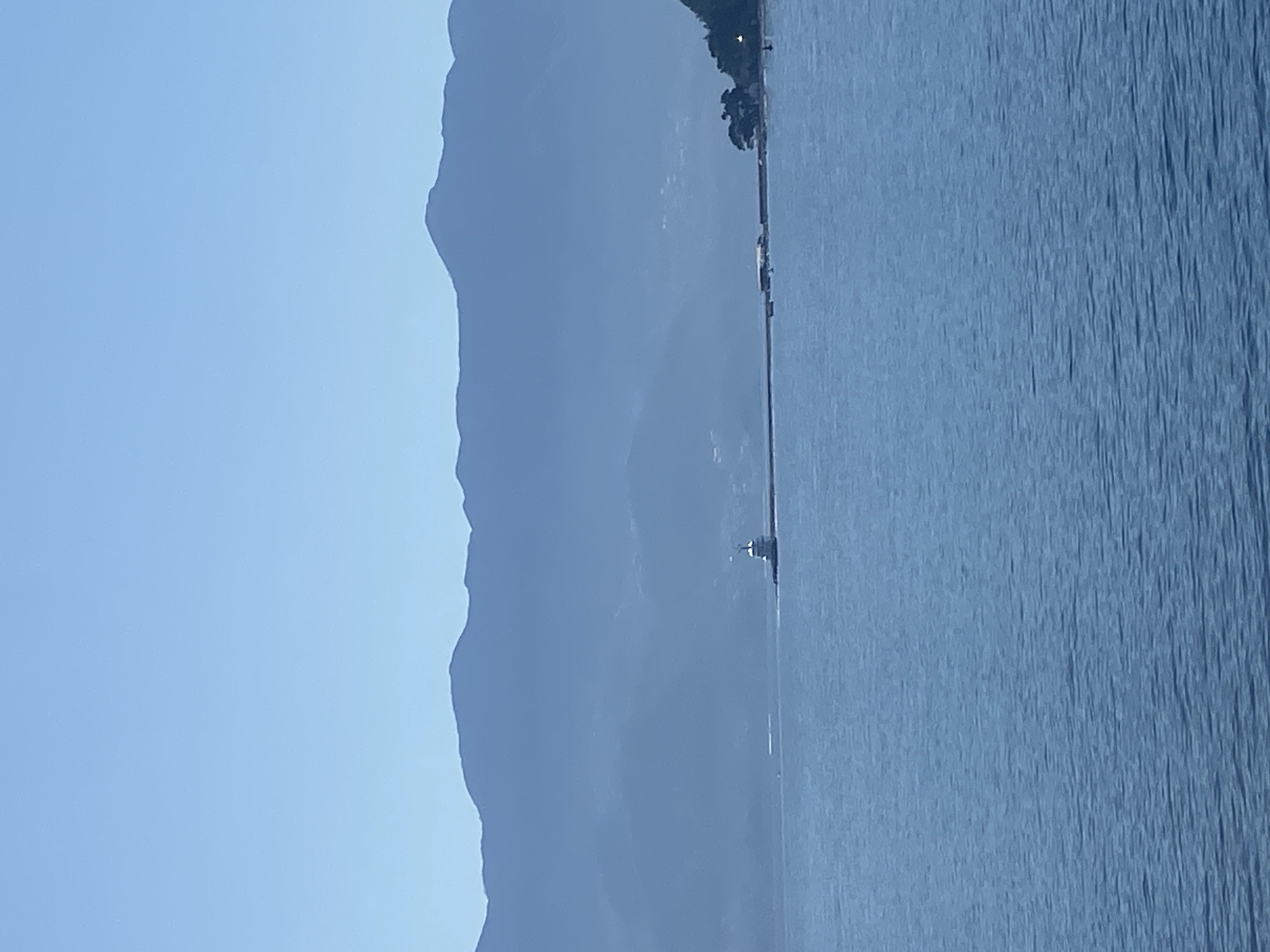 The image shows a waterscape with a mountainous backdrop, likely taken in Budva, Montenegro. On the right side, the water surface is visible. A land formation is observed to the left, with visible elevation changes indicating mountainous terrain. A boat can be seen, small in the frame, suggesting a vastness in scale. The sky occupies the upper part of the image, suggesting daytime conditions.