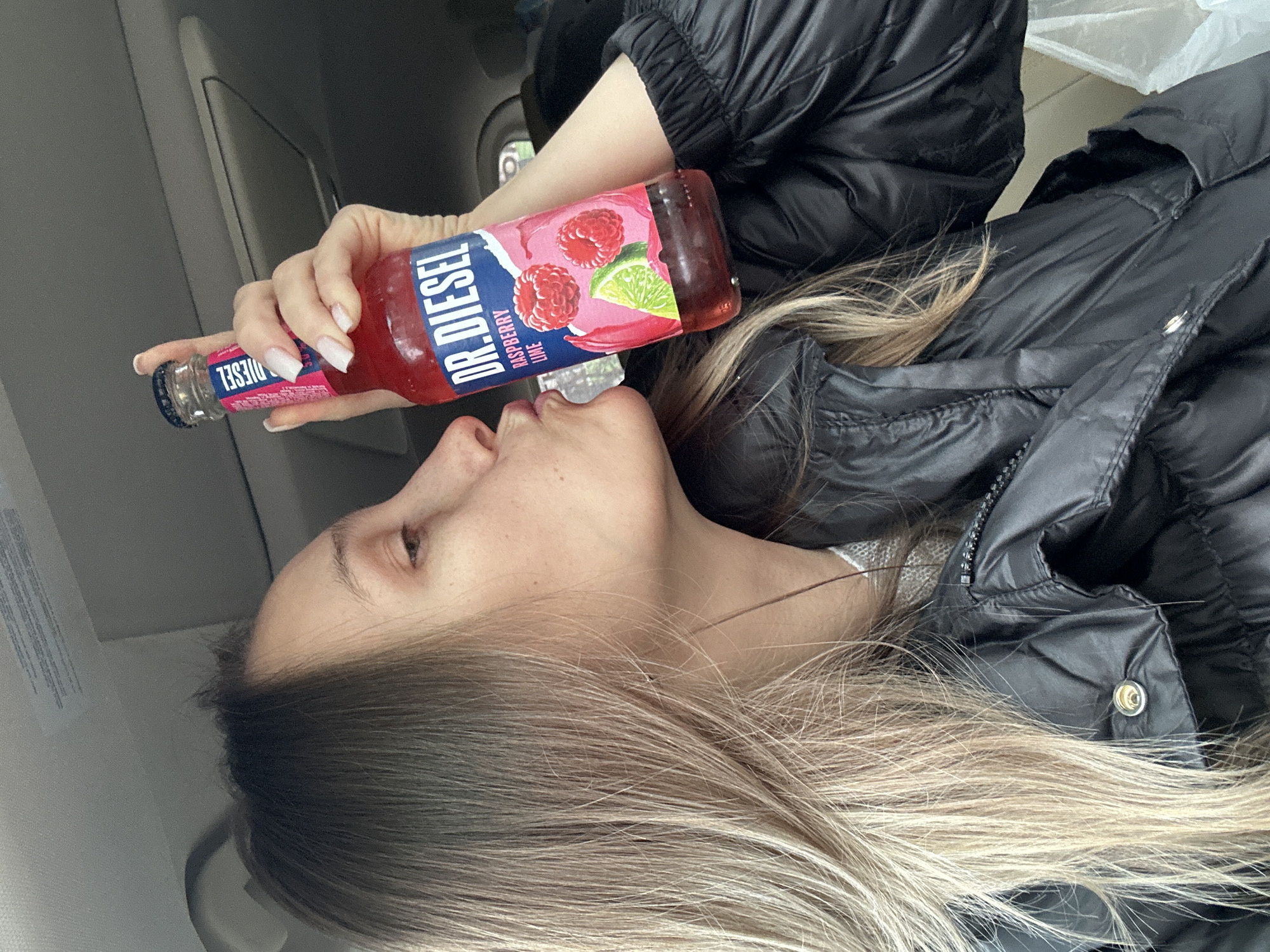 A woman is drinking from a bottle of "Dr. Diesel Raspberry Lime" flavored soda. She is wearing a black puffy jacket and has blonde hair. The location appears to be inside a vehicle. There is no indication that the location is in the city of Myskhako, Russia.