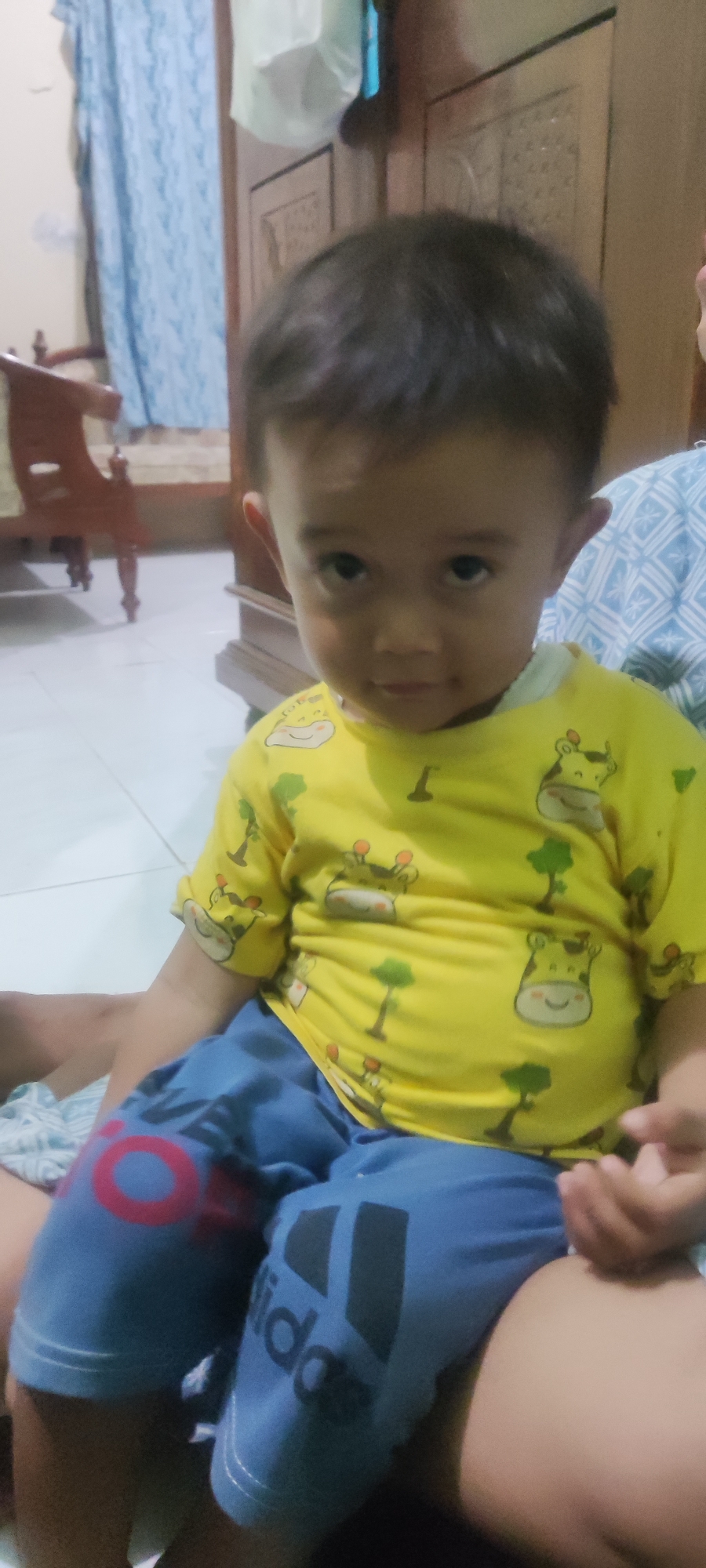 A young child with dark hair and large, dark eyes is seated, wearing a yellow t-shirt with a cartoon giraffe pattern and blue pants with Adidas branding on the left leg. The child is positioned indoors, likely in a residential setting in Gebang, Indonesia, with a wooden wardrobe visible in the background, along with a patterned blue textile, possibly a sofa or blanket. The floor appears to be tiled. The lighting suggests an indoor environment, possibly during the day. The child is looking towards the camera with a neutral expression.