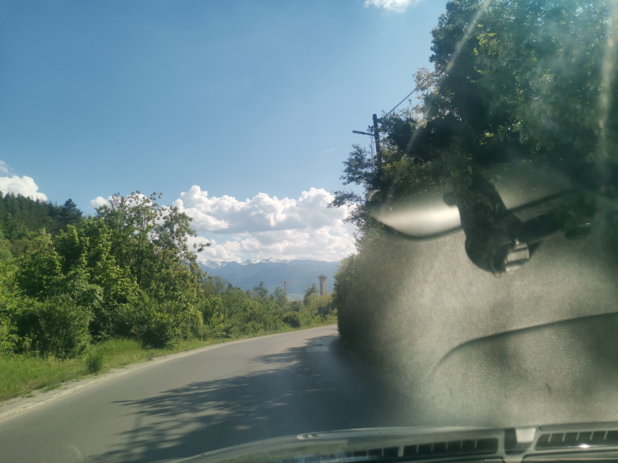 The image shows a road in Momin Prohod, Bulgaria. The view is from inside a car looking through the windshield. The road is paved and curves slightly to the right. On the left side of the road is green vegetation, and beyond that, there are mountains in the distance, partially covered in snow. There are cumulus clouds in the sky. On the right side of the road, there is also vegetation. There's a utility pole with wires visible. In the distance, a tower crane and tall structure can be seen. The interior of the car is visible at the bottom of the frame, with the dashboard and windshield wiper arm prominent. A rearview mirror is visible attached to the windshield. Sunlight is reflected on the windshield.
