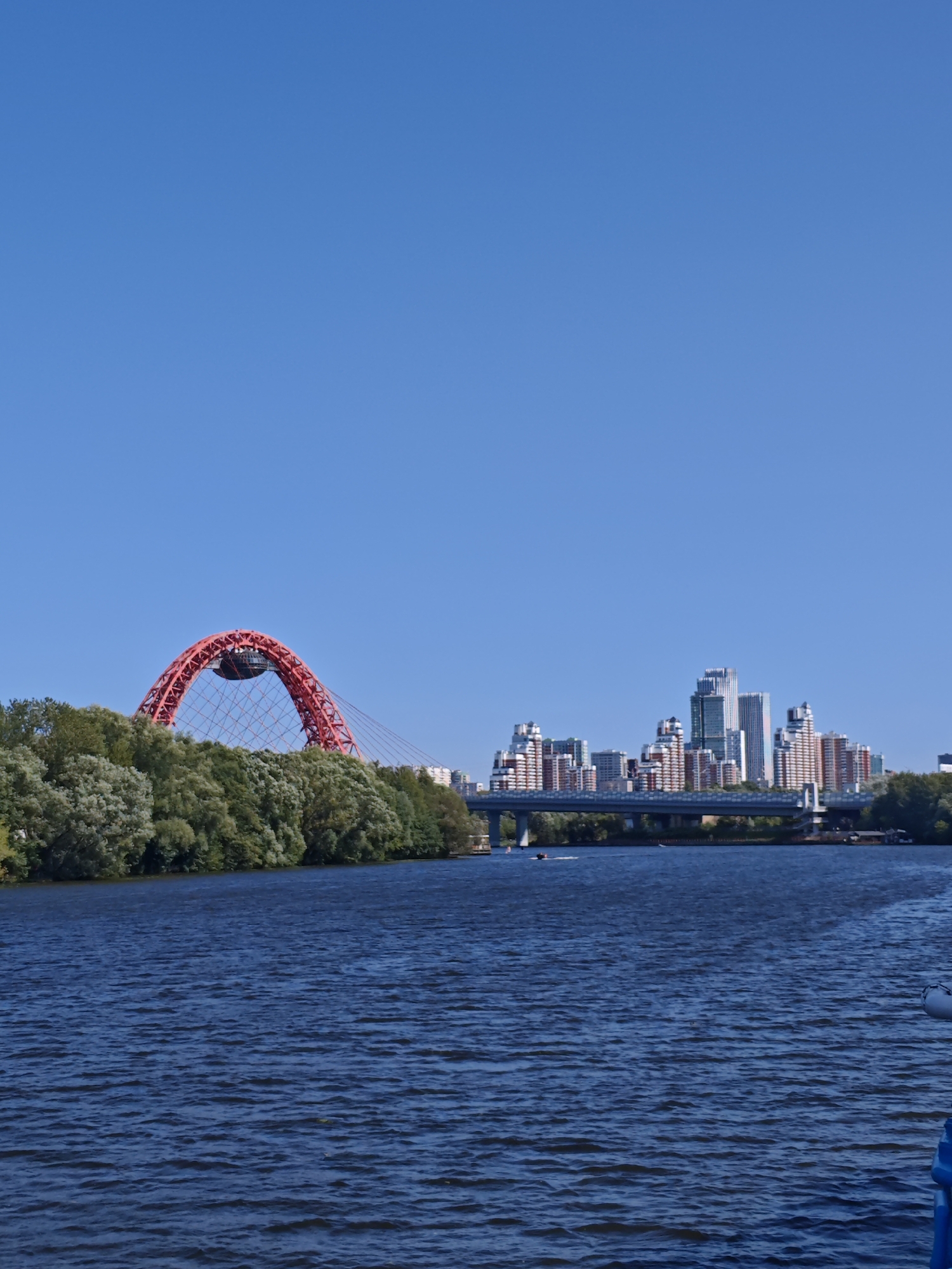 This is a photograph taken from the water in Moscow, Russia, showing a cityscape with a distinctive red arch bridge and modern buildings under a clear blue sky. The foreground is dominated by the dark blue water of a river, with ripples indicating some movement.
On the left side of the image, the prominent feature is a bright red suspension bridge with a large arch, known as the Zhivopisny Bridge. It spans across the river, with its intricate metal structure visible within the arch. Behind the bridge, a dense line of lush green trees forms a natural border along the riverbank.
In the background, a cluster of modern buildings, including high-rises and apartment complexes with various architectural styles and colors (predominantly white, beige, and some green), creates a city skyline. A road bridge crosses the river in front of these buildings.
The water surface reflects the blue of the sky. A few small boats are visible on the water, suggesting recreational activity. The overall scene depicts a sunny day in an urban environment where nature and modern architecture coexist along a waterway.