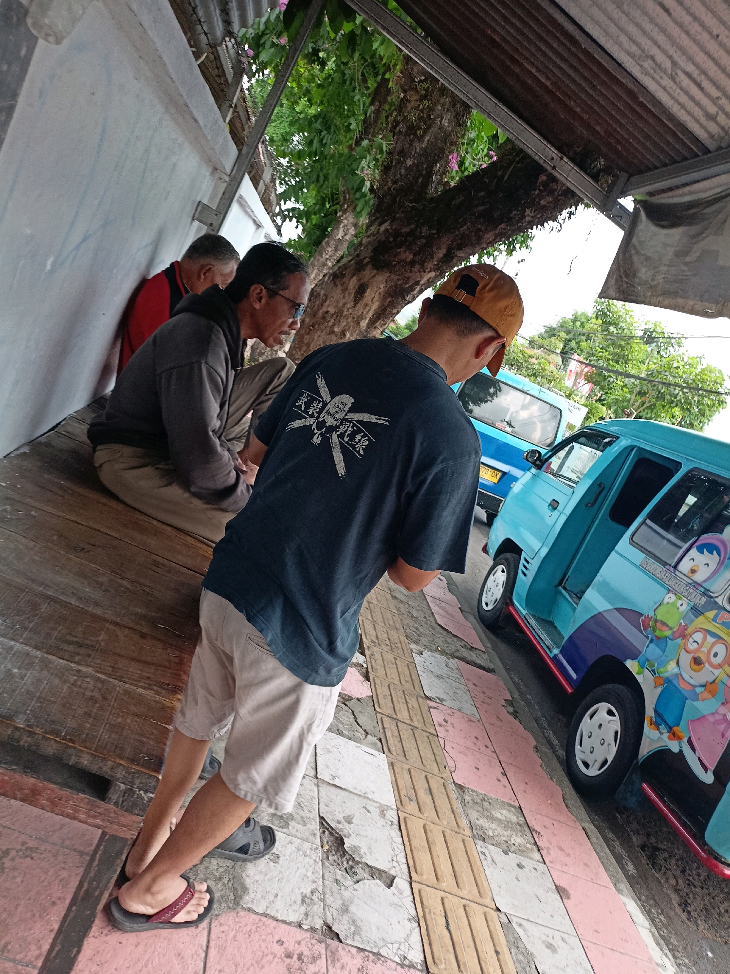 The image shows three men in the city of Magelang, Indonesia. Two men are sitting on a wooden bench against a white wall, while a third man stands facing away from the camera, looking at two light blue vans. The van in the foreground is decorated with cartoon characters. The scene appears to be outdoors, possibly in a shaded area under a roof or awning, with trees and foliage visible in the background. The ground is a combination of wooden planks and paved walkway with textured tiles.