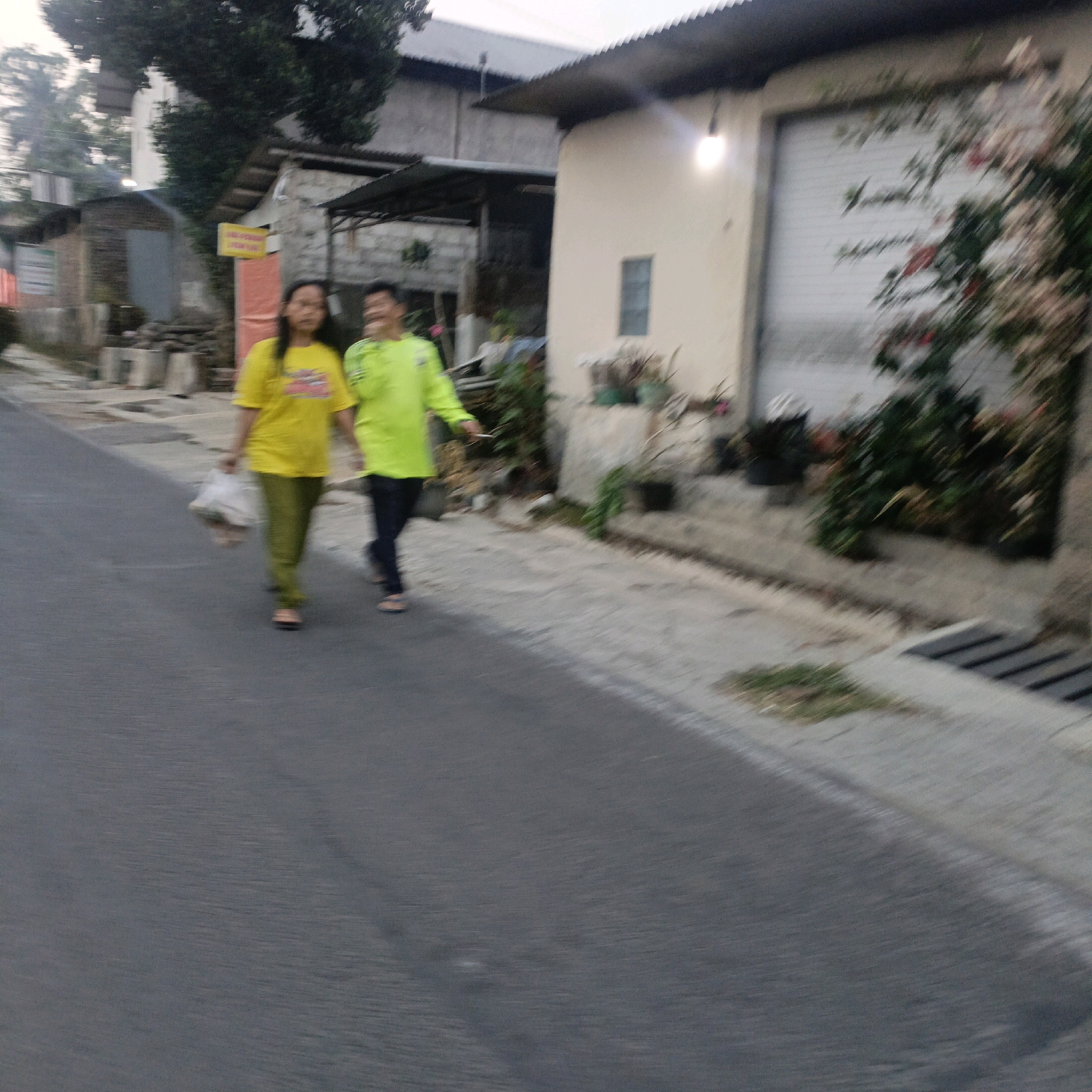 Two people are walking on an asphalt road in front of a building. The person on the left is wearing a yellow t-shirt and green pants. They are carrying a white plastic bag in their left hand. The person on the right is wearing a green long-sleeved shirt and dark pants. They are walking towards the right of the frame. In the background, there is a building with a large roller door and some greenery. The scene appears to be in an urban or semi-urban area. The image quality is low, with motion blur affecting clarity.