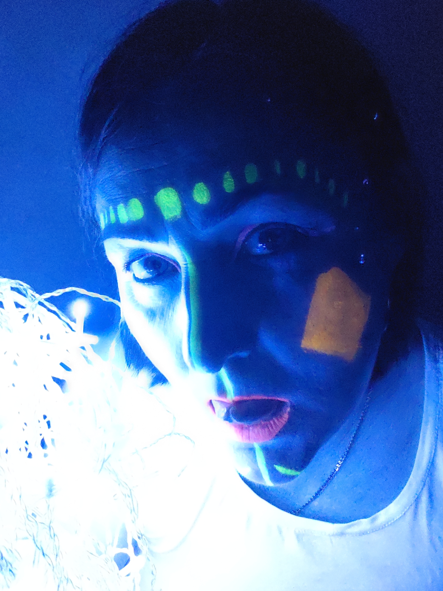 A person is depicted in a close-up shot, appearing to be under ultraviolet (UV) or blacklight illumination. The individual has neon-colored face paint or makeup in green, yellow, and pink. The person holds a string of bright white lights, which are partially obscuring the lower left of the frame. The background is dark and undefined. The setting is not identifiable as Markovskii, Russia, based on the provided image alone. No further information on the location is available from the image.