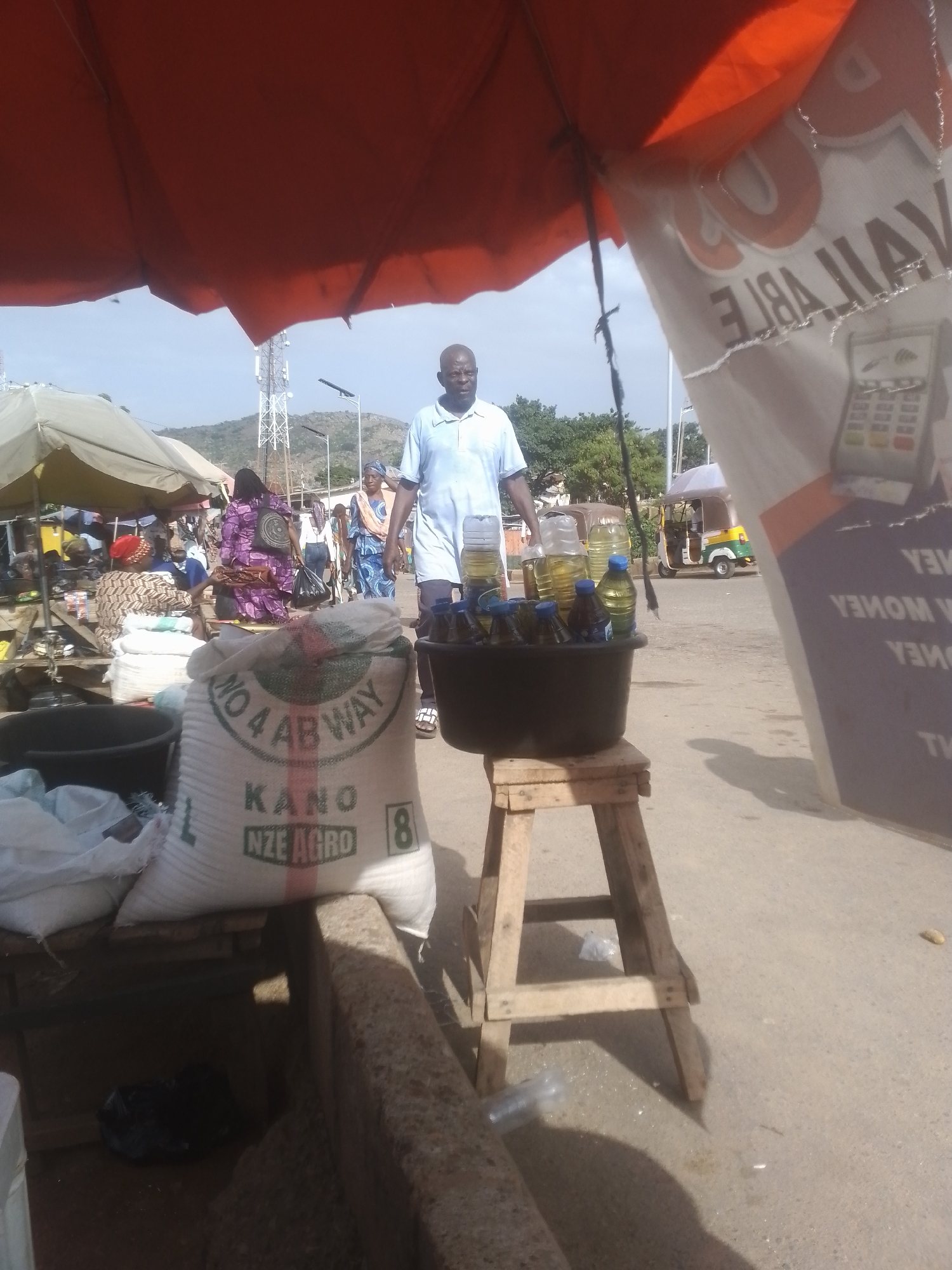 An eye-level, medium outdoor shot shows a man wearing a blue shirt and white shorts standing in what appears to be a market stall in front of a white and orange awning. To the left, a woman in a purple and black patterned dress is visible in the background, and to the right, a large white bag with text that reads "KANO NZE AGRO" is prominently displayed. A black basin filled with bottles of cooking oil sits on a wooden stool in the foreground. In the background, a telecommunications tower and several vehicles, including an auto-rickshaw, are visible under a bright sky. The ground is a light brown, dusty surface.