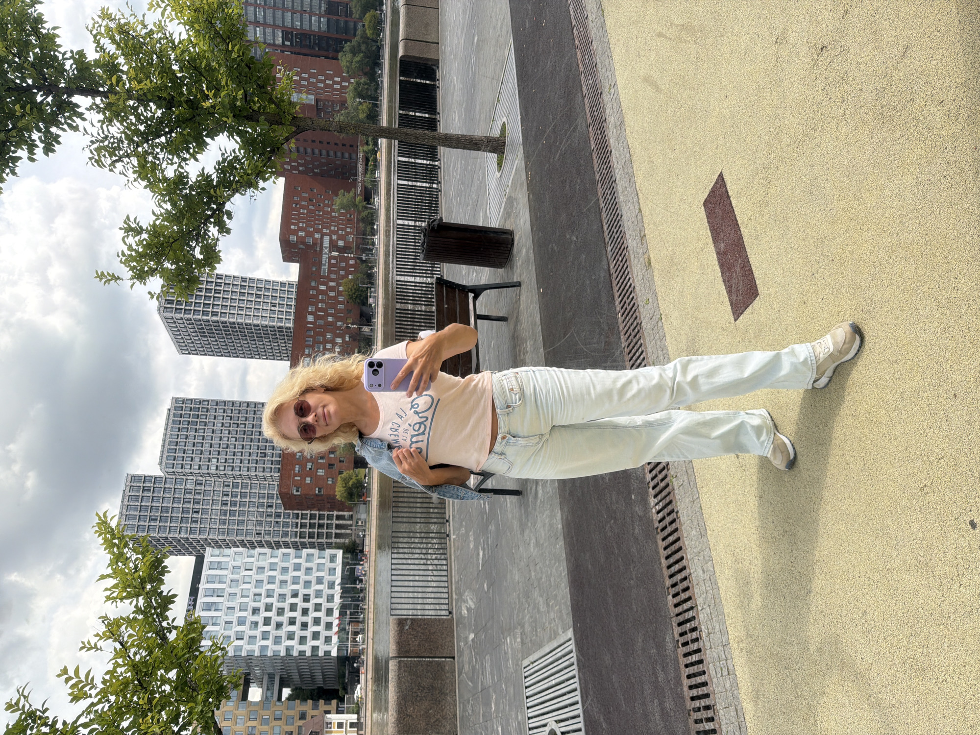 A woman stands on a yellow paved walkway, taking a selfie with a purple smartphone. She wears a light pink t-shirt with blue text, light blue jeans, and beige sneakers. A denim jacket is slung over her left shoulder. She has blonde hair and is wearing sunglasses. Behind her, a river is visible with a metal fence along its edge. Further back, there are several modern, multi-story buildings with various architectural styles, some with many windows and some with brick facades. Trees with green foliage are present in the foreground and background. The sky is cloudy. The location appears to be an urban waterfront area, likely in Moscow, Russia, possibly along the Moskva River, given the architectural style and waterfront development common in the city.