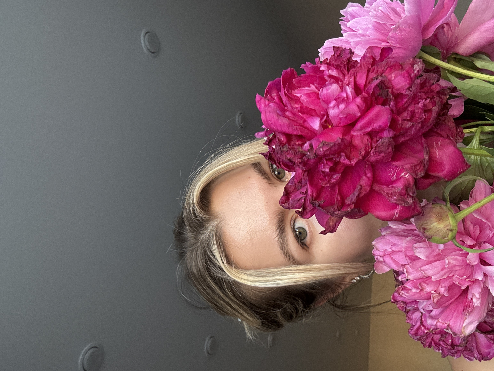 In this close-up shot, a young woman is partially obscured by a vibrant bouquet of pink peonies. Her visible eye, with its green iris, looks directly at the camera. She has light brown hair with blonde highlights, pulled back loosely. The background is a neutral gray ceiling with circular light fixtures. The peonies, in varying shades of pink and magenta, dominate the foreground, with some petals appearing slightly wilted, suggesting the flowers are not freshly cut. The lighting is soft, coming from the front and to the left.