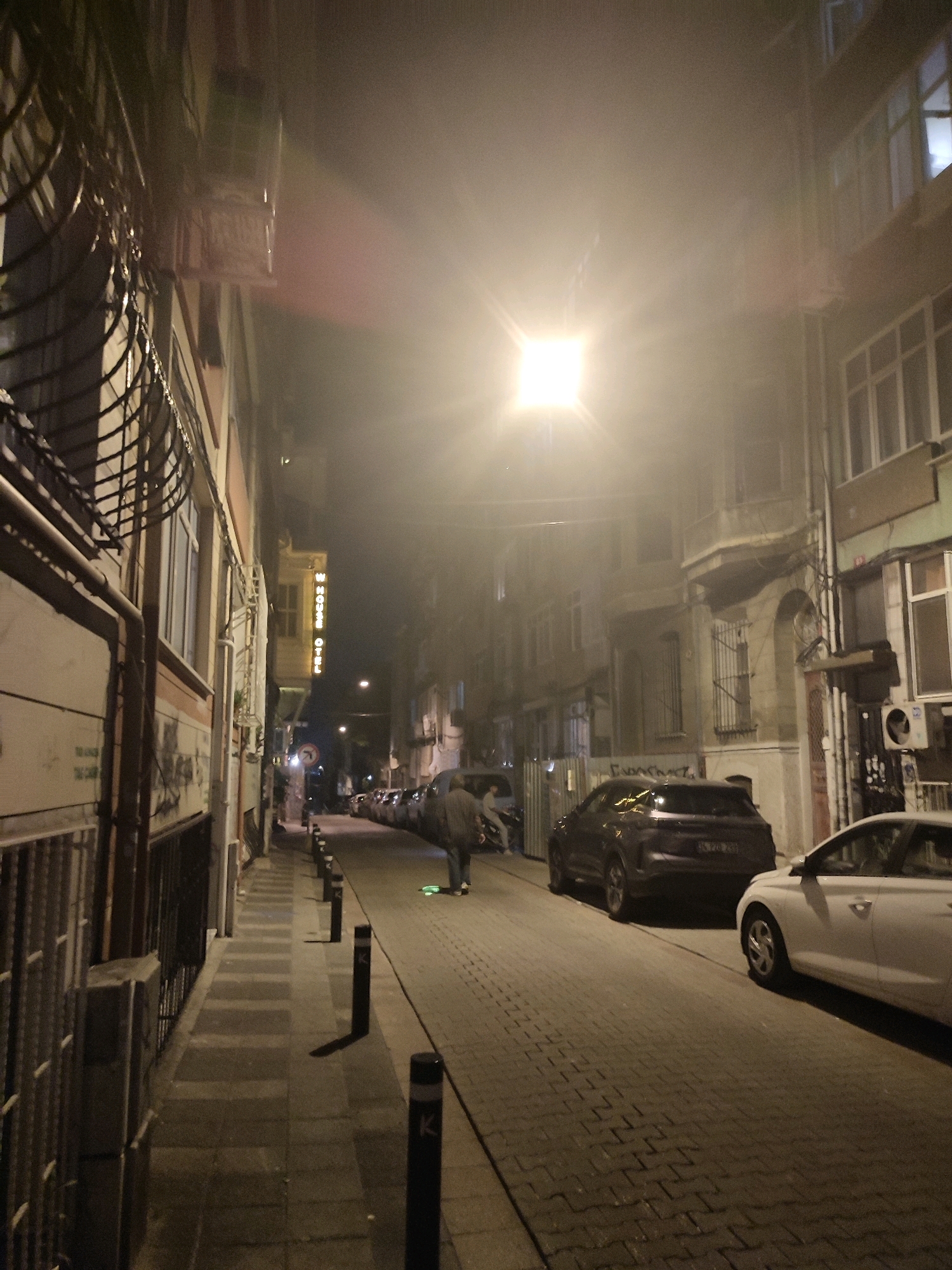 The image depicts a nighttime street scene in Kadıköy, Turkey, characterized by foggy or misty conditions. A bright, square light source illuminates the upper portion of the frame, creating a lens flare. The street is paved with cobblestones and lined with buildings on both sides. On the left, a building features barred windows and a drainpipe. A yellow neon sign with vertical text, possibly indicating "HOTEL," is visible further down the street on the right.
Several parked cars are present, including a dark SUV and a white sedan in the foreground. A person is walking away from the camera on the sidewalk to the right, appearing to be on a skateboard or similar device, with a faint green glow visible beneath them. The overall atmosphere is subdued and somewhat mysterious due to the low light and fog.