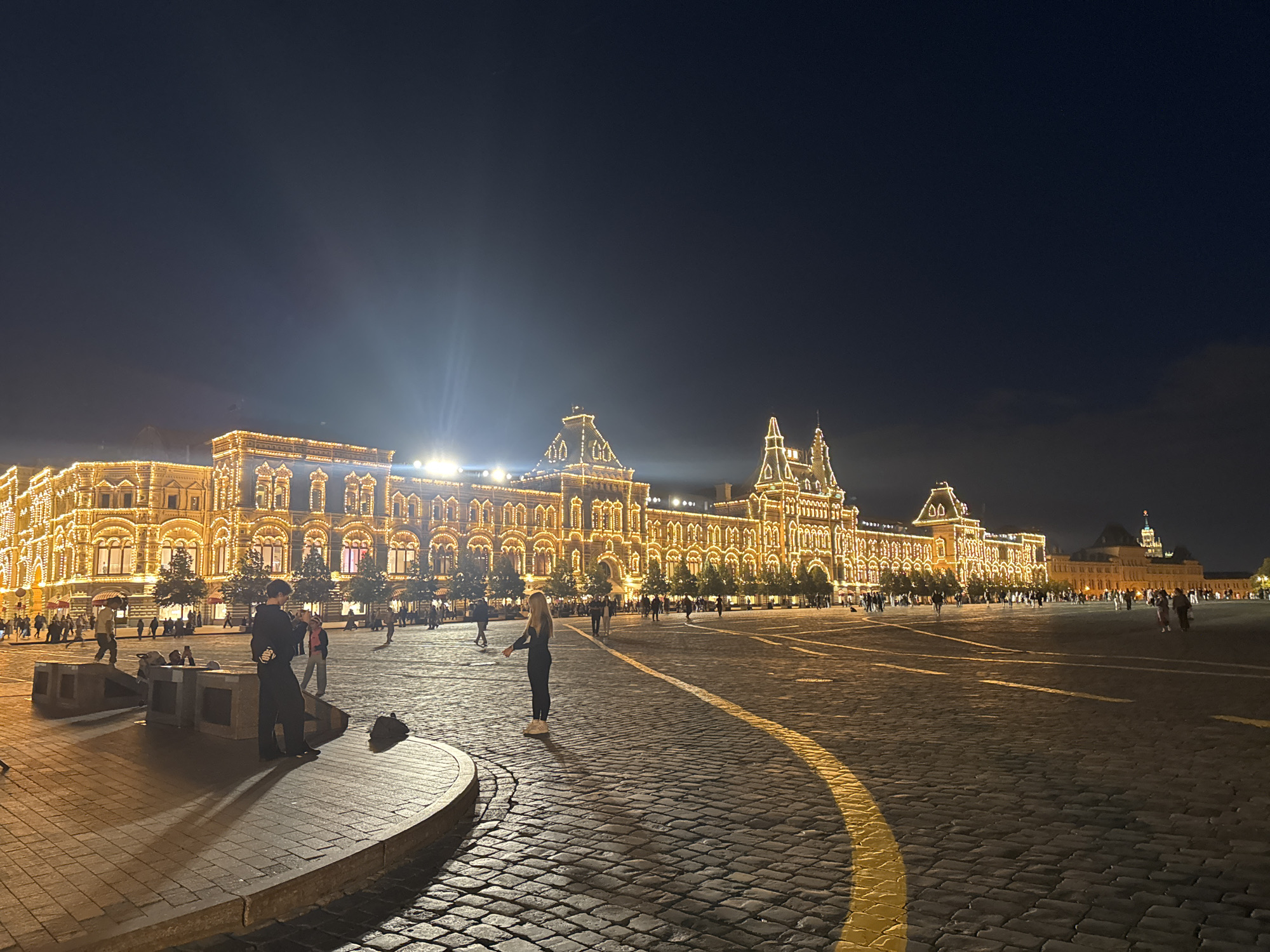 The image depicts a nighttime scene in Red Square, Moscow, Russia. The GUM department store is prominently featured, illuminated by thousands of golden lights outlining its architecture. The cobblestone pavement of the square is visible, with a yellow line marking a curved path. Several individuals are present in the foreground and middle ground, engaged in various activities such as walking, standing, and posing for photos. Trees are sparsely planted along the front of the GUM building, also adorned with lights. The sky is dark, indicating nighttime, with some faint clouds visible. The overall impression is one of a bustling urban square at night, with a focus on the illuminated facade of a historic building.