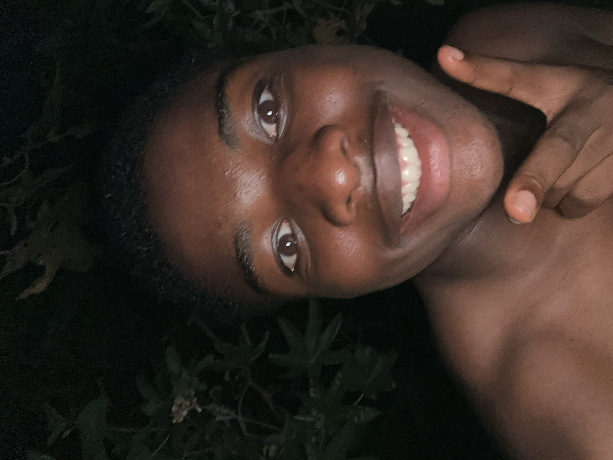 An individual with dark skin smiles brightly at the camera in a close-up, selfie-style photograph. The person is lying down, with their head tilted, and their gaze directed upwards, meeting the viewer's eyes. A wide, open-mouthed smile reveals a full set of white teeth, conveying an emotion of happiness or amusement. Their right hand is raised, with the index finger pointing upwards.
The background is dark and appears to be an outdoor, natural setting, possibly a garden or area with foliage, indicated by faint green shapes of leaves and plants. This suggests the image was taken at night or in very low light conditions. The person's upper body is bare, showing smooth, dark skin on the shoulder and chest. The lighting, while dim overall, highlights the person's face and smile. The scene evokes a feeling of warmth and joy within a dark, natural environment. The location context is Bar Gada, Nigeria. No visible text is present in the image.