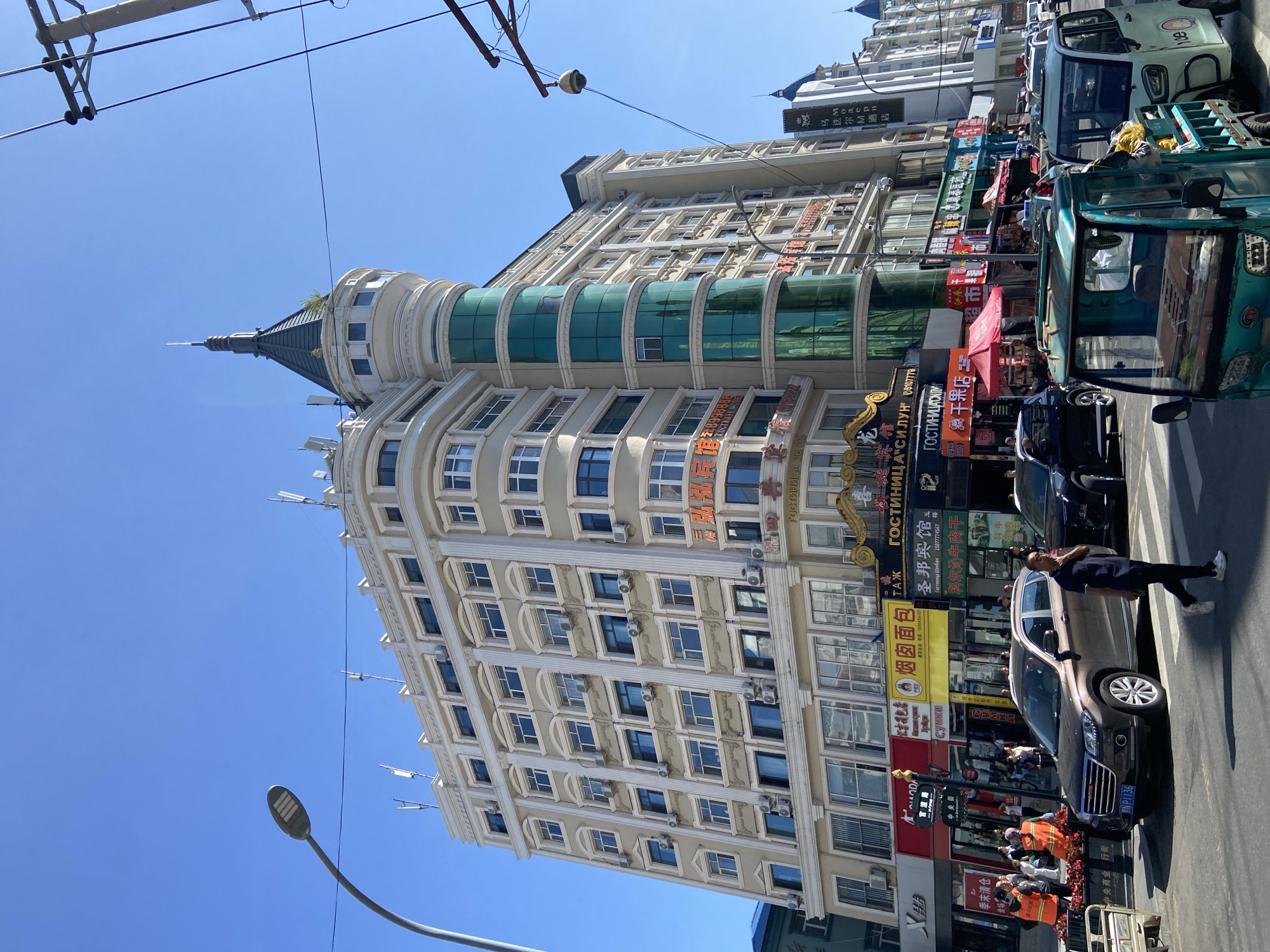 In Xing'an Subdistrict, China, a multistory building with a distinctive turret stands prominently on a sunny day. The building features a beige facade with numerous windows, many equipped with air conditioning units. The turret, a pointed structure with a dark green roof and glass windows, is integrated into the building's design. Street level is bustling with activity, featuring various shops with colorful signage in Chinese and Russian. A street lamp on a curved pole is visible to the left, and power lines crisscross the sky. Pedestrians are walking on the sidewalk, and several vehicles are present, including a brown SUV, a dark sedan, and two three-wheeled vehicles. One of the three-wheeled vehicles is light blue and appears to be a cargo type, while the other is light green and has "ICE" written on its side. People in orange vests are also visible near the shops. The scene captures a typical urban streetscape with commercial activity and everyday life.