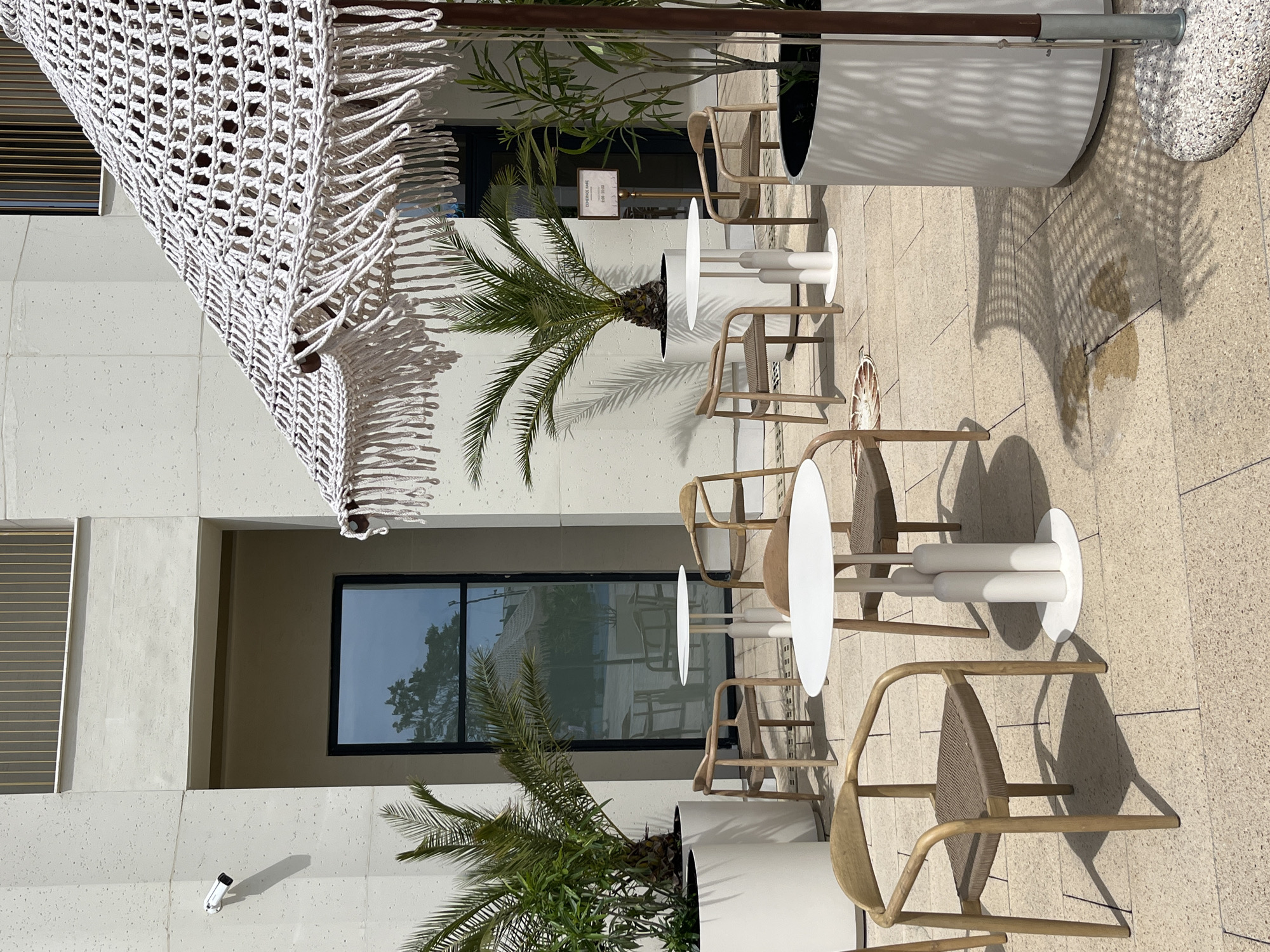 The image shows an outdoor seating area, likely a cafe or restaurant, in an urban environment. The scene features several white round tables surrounded by light wood chairs with woven seats. Each table is flanked by two chairs. The tables have distinctive white bases consisting of stacked cylinders. Large white planters containing palm trees and other greenery are placed near the seating arrangements. An awning or sunshade with a decorative white macrame fringe is visible in the upper left corner of the image. The background shows a modern building with rectangular windows, some reflecting the outdoor scenery. The flooring appears to be light-colored stone tiles. Shadows cast by the objects indicate bright sunlight. A sign with text is positioned near one of the planters. The overall impression is that of a stylish and inviting outdoor space. It is impossible to confirm the location is in Sirius, Russia.