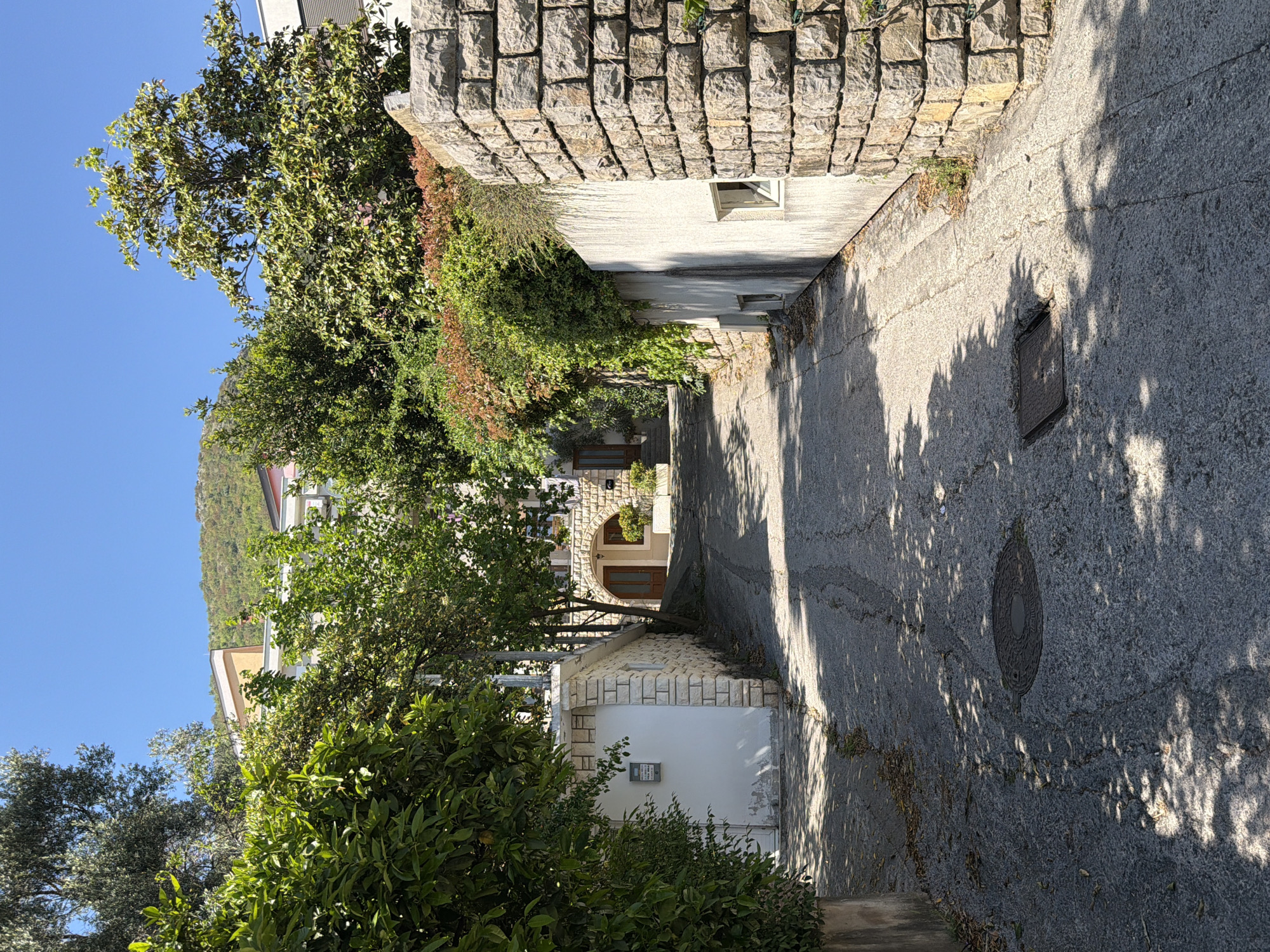 The image displays a sunlit, narrow alleyway or path in Nerin, Montenegro, leading gently downwards between structures and dense vegetation. The path, made of concrete or asphalt, shows signs of cracks and wear. Long, distinct shadows from trees fall across its surface, indicating a bright, sunny day, likely during the morning or late afternoon, under a clear blue sky.
On the right side of the path, a prominent stone wall is visible, partially covered with green and reddish-brown vines and bushes. Below this wall is a light-colored building with a small, rectangular window set into its facade. Further along the path on this side, another building features a distinctive arched entrance with a brown wooden door.
The left side of the path is bordered by lush green trees and shrubs. Partially obscured by this foliage is a white building, to which a smaller, stone-accented structure is attached. A grey rectangular box, possibly an intercom or mailbox, is mounted on the white wall of this structure, bearing the visible text "D. KNEZEVIC" and "20".
In the background, beyond the buildings and dense greenery, a verdant, forested hillside rises against the cloudless blue sky. Two manhole covers are embedded in the path: one square, closer to the foreground, and another round/oval-shaped one further down. No people or specific activities are depicted in the scene.